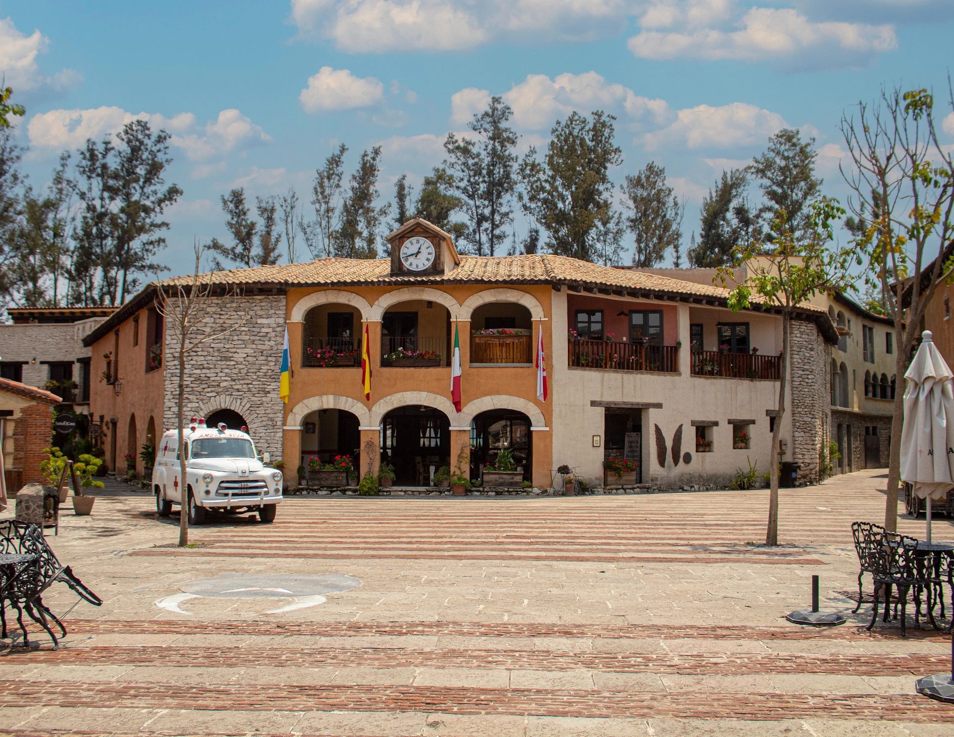 A two-story building in a plaza, beige and stone facade, clock on top, flags, and an old truck.