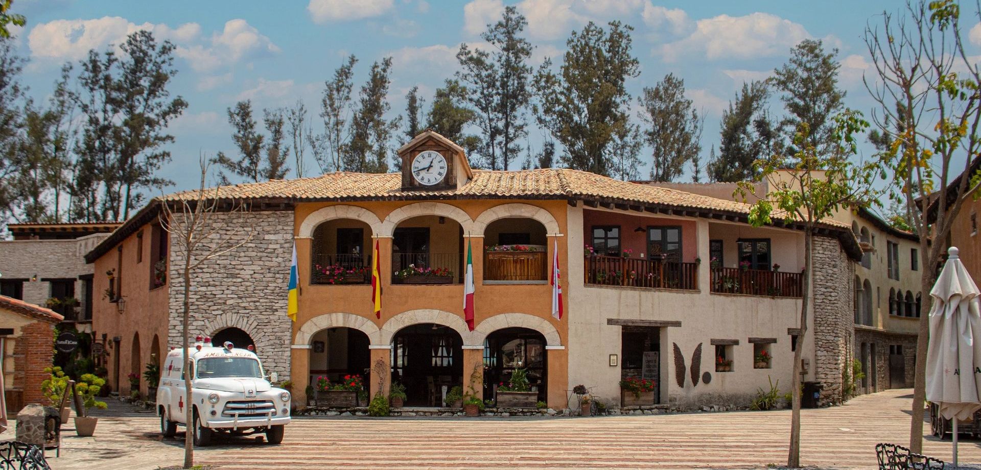 A two-story building with arches and flags. A clock is above the arches, with a white truck parked in front.