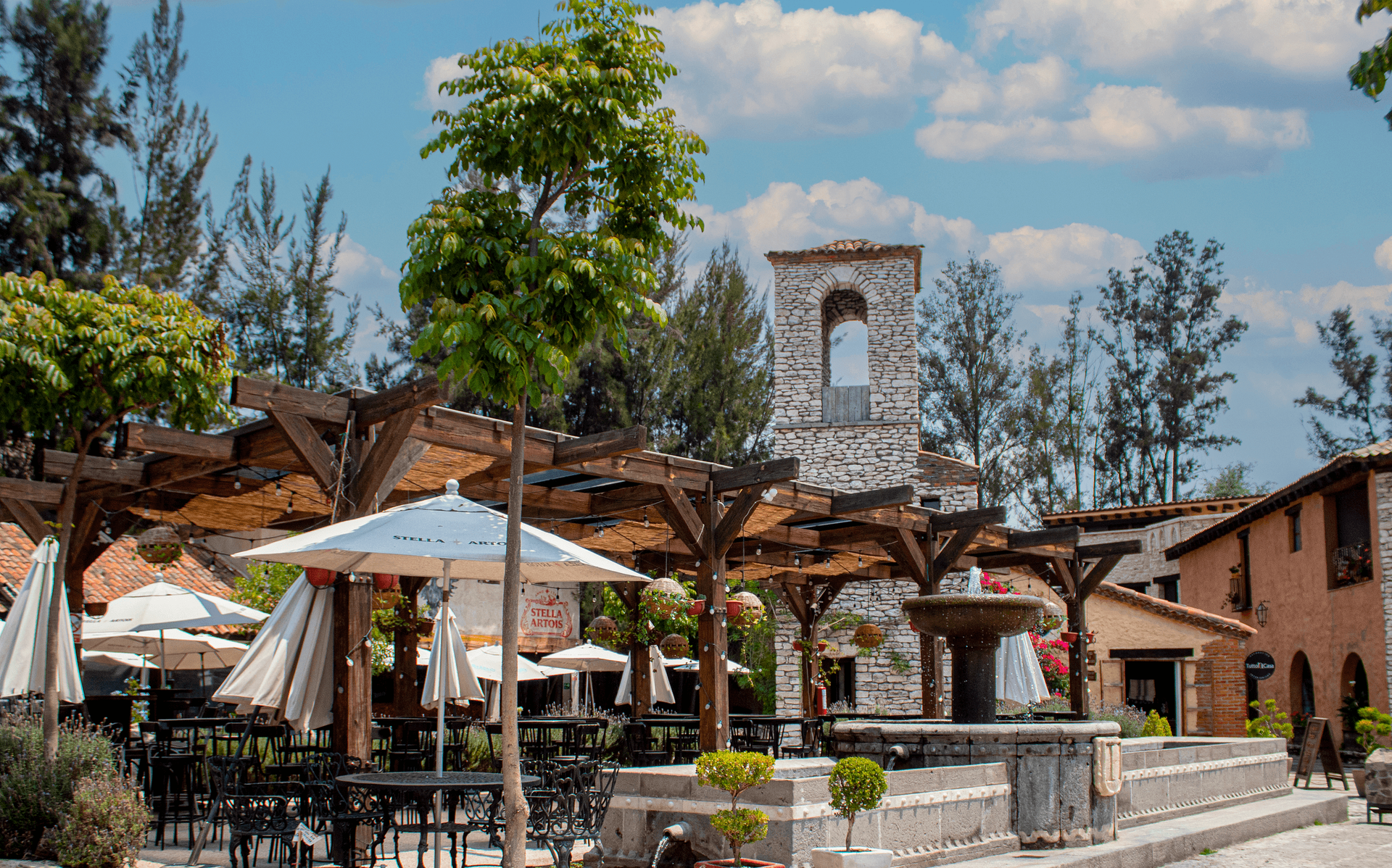 Outdoor plaza with restaurants, umbrellas, fountain, and stone tower under a blue sky.