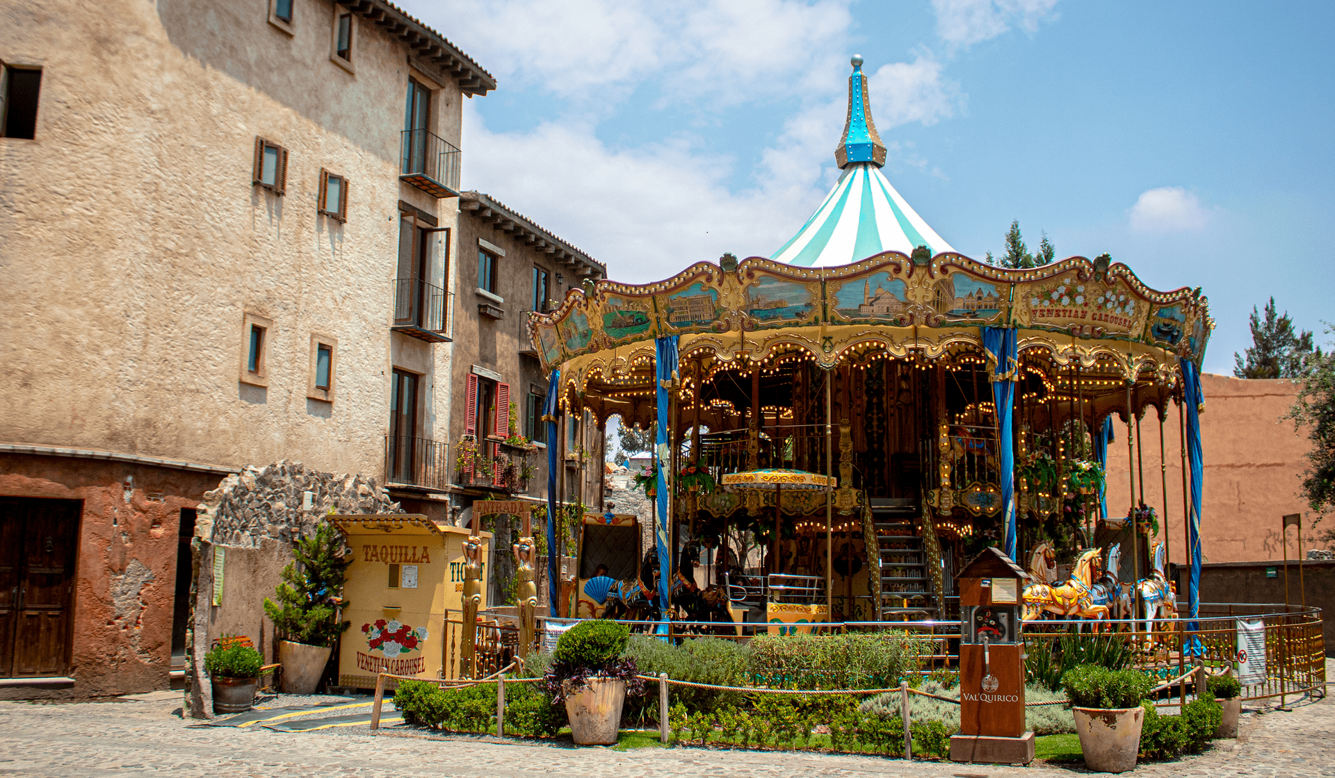 A vintage carousel with gold accents and blue/green canopy stands next to a stone building in a plaza.