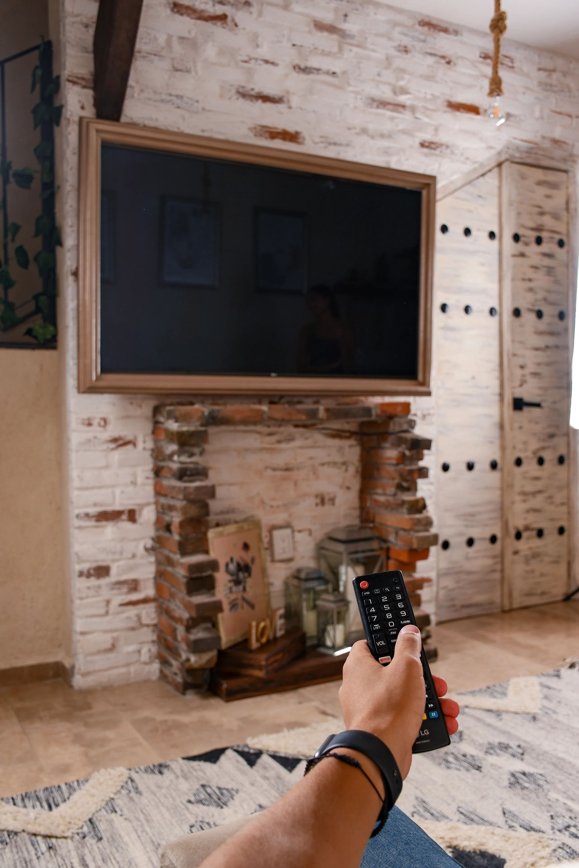 Person holding remote, watching a TV mounted above a brick fireplace in a rustic room.
