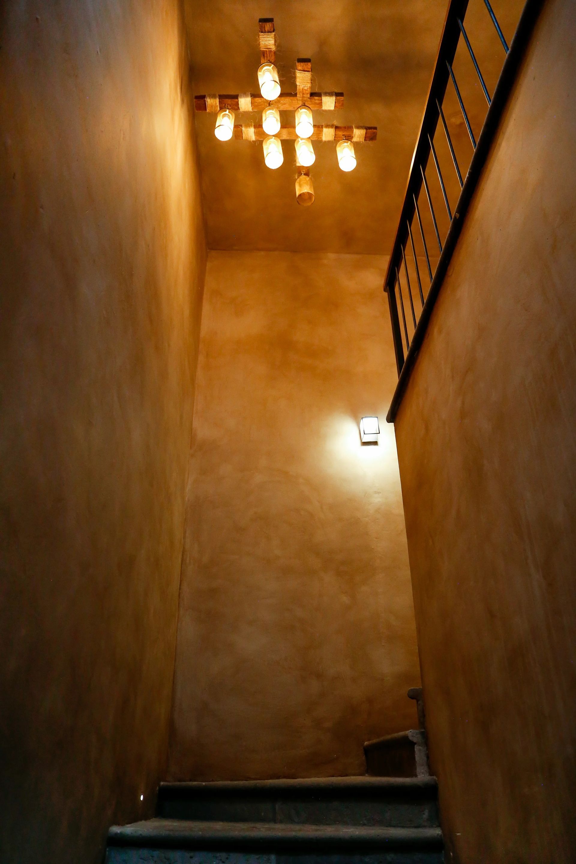 Stairwell with textured brown walls, lit by a unique light fixture and a wall-mounted light. Black railing.