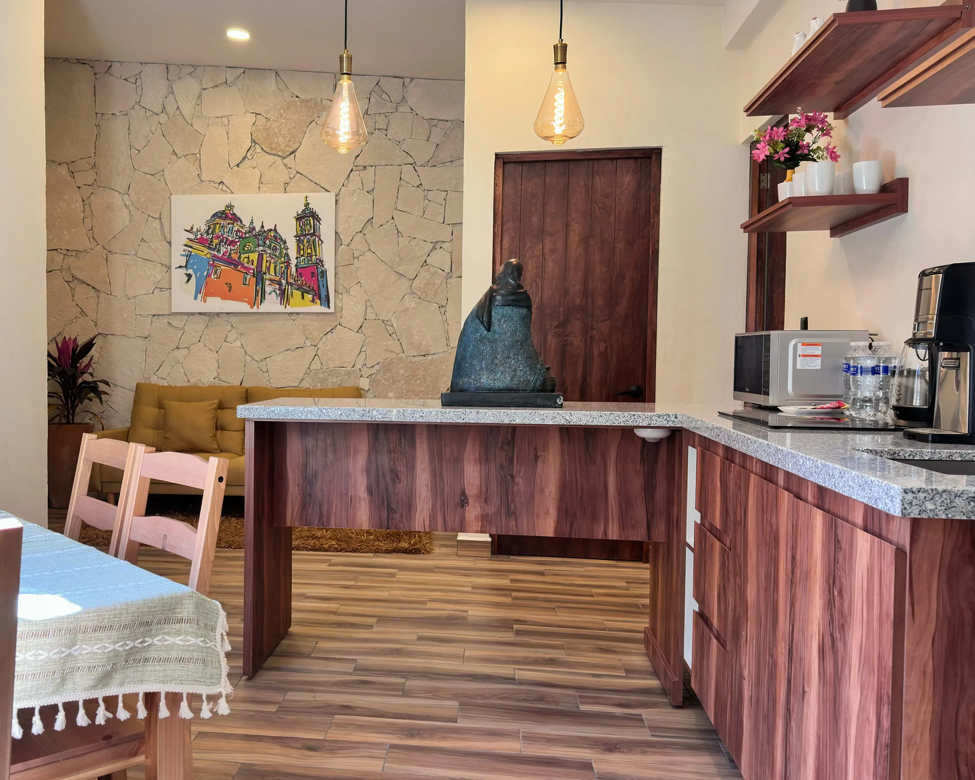 Una cocina con gabinetes de madera, encimera de piedra y mesa de comedor. Un cuadro cuelga de una pared texturizada.