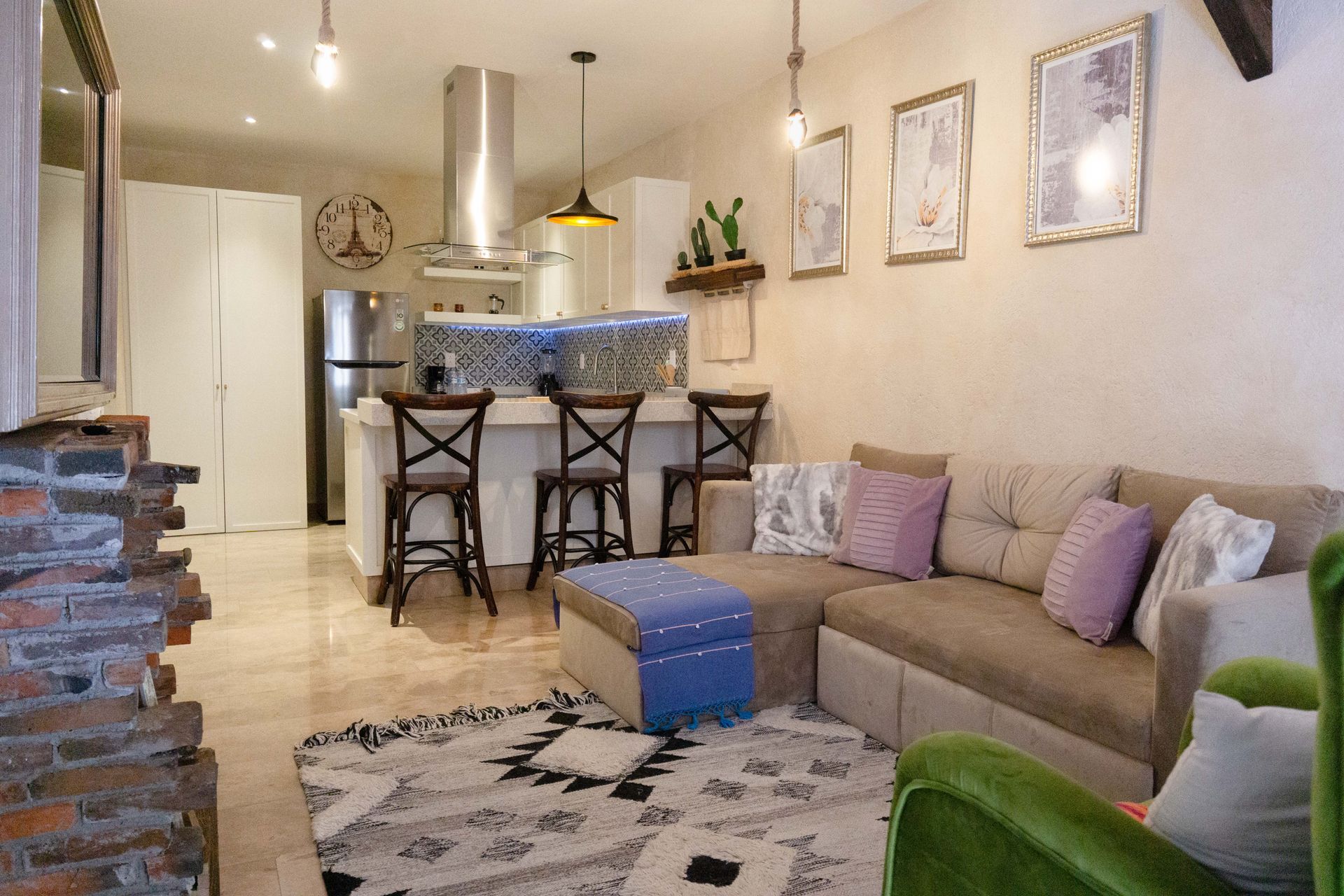 Cozy living room with beige sofa, blue ottoman, bar stools, and open kitchen in the background