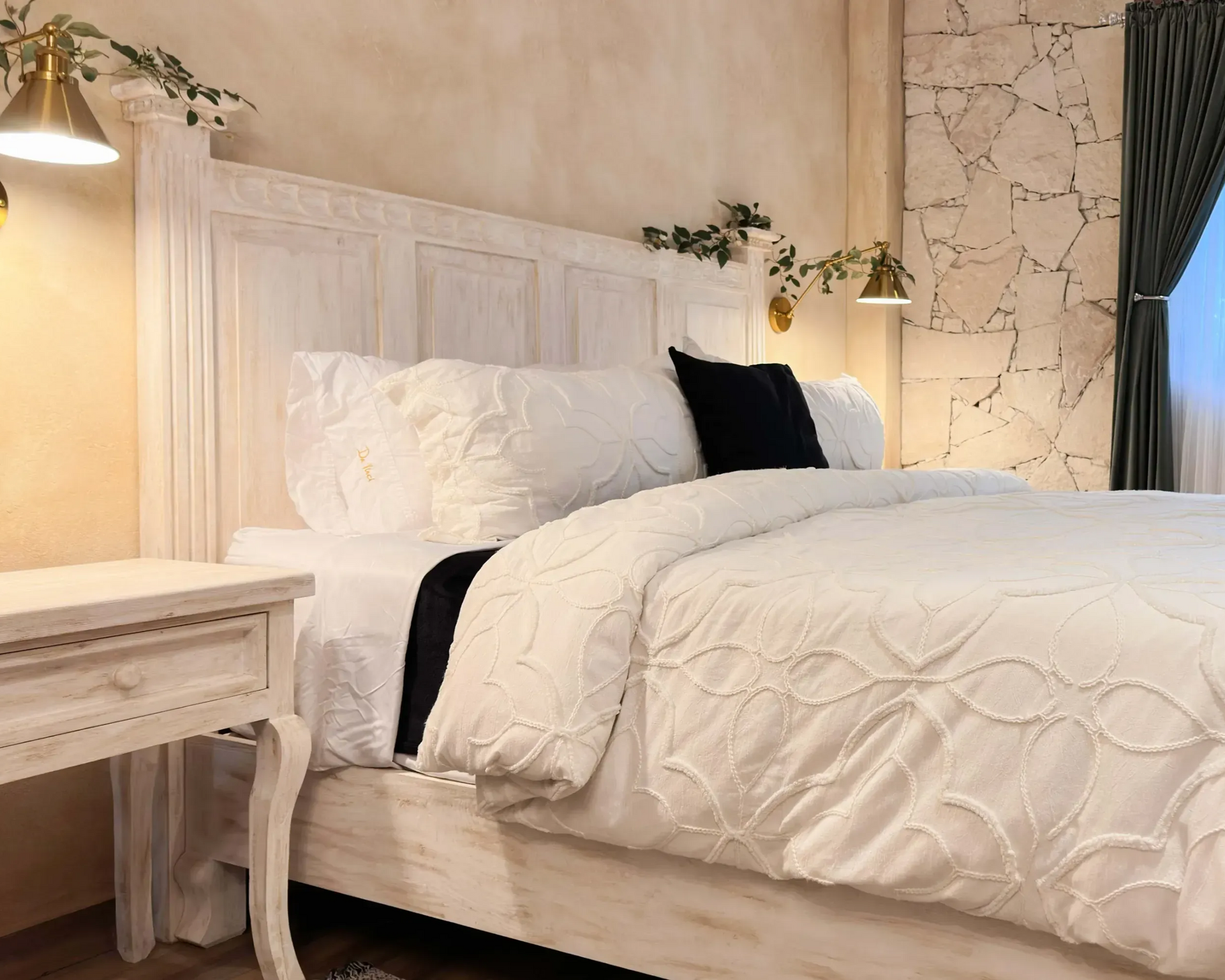Dormitorio blanco con cama, mesita de noche, luces de latón y vegetación decorativa.