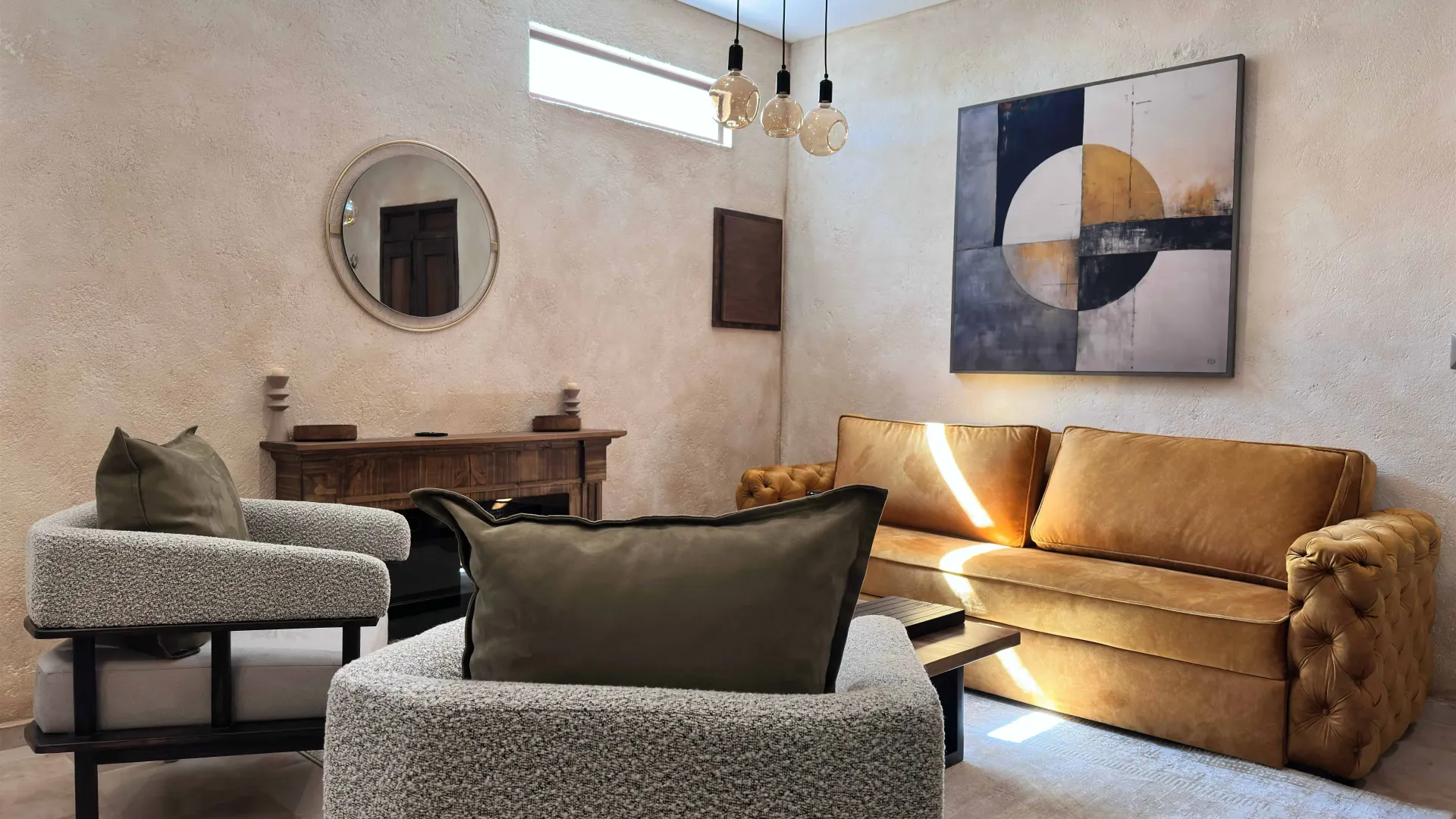 Living room with mustard sofa, gray chairs, art, and round mirror on textured walls.