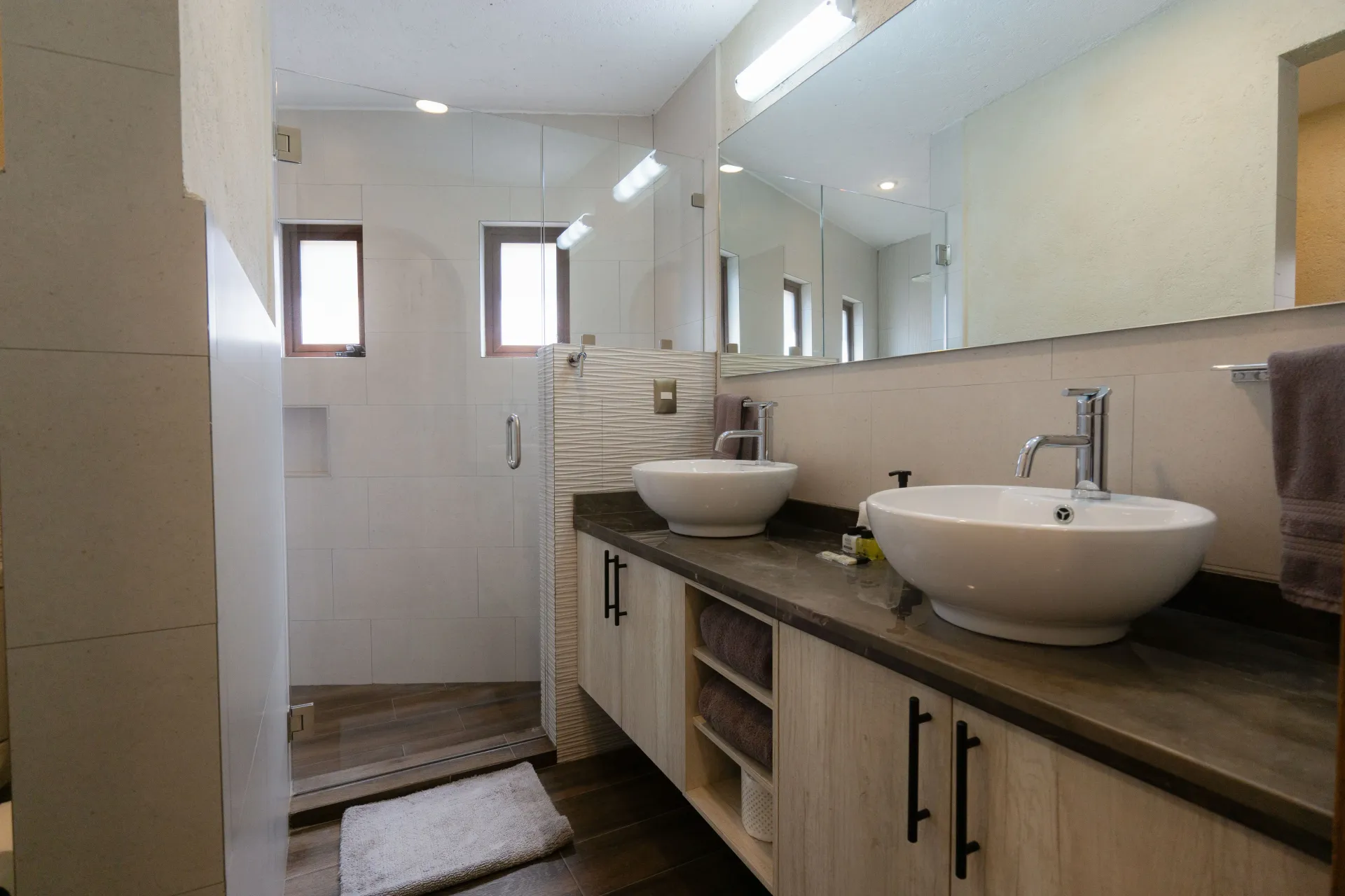 Bathroom with two sinks, large mirror, shower, light-colored cabinets, and small windows.