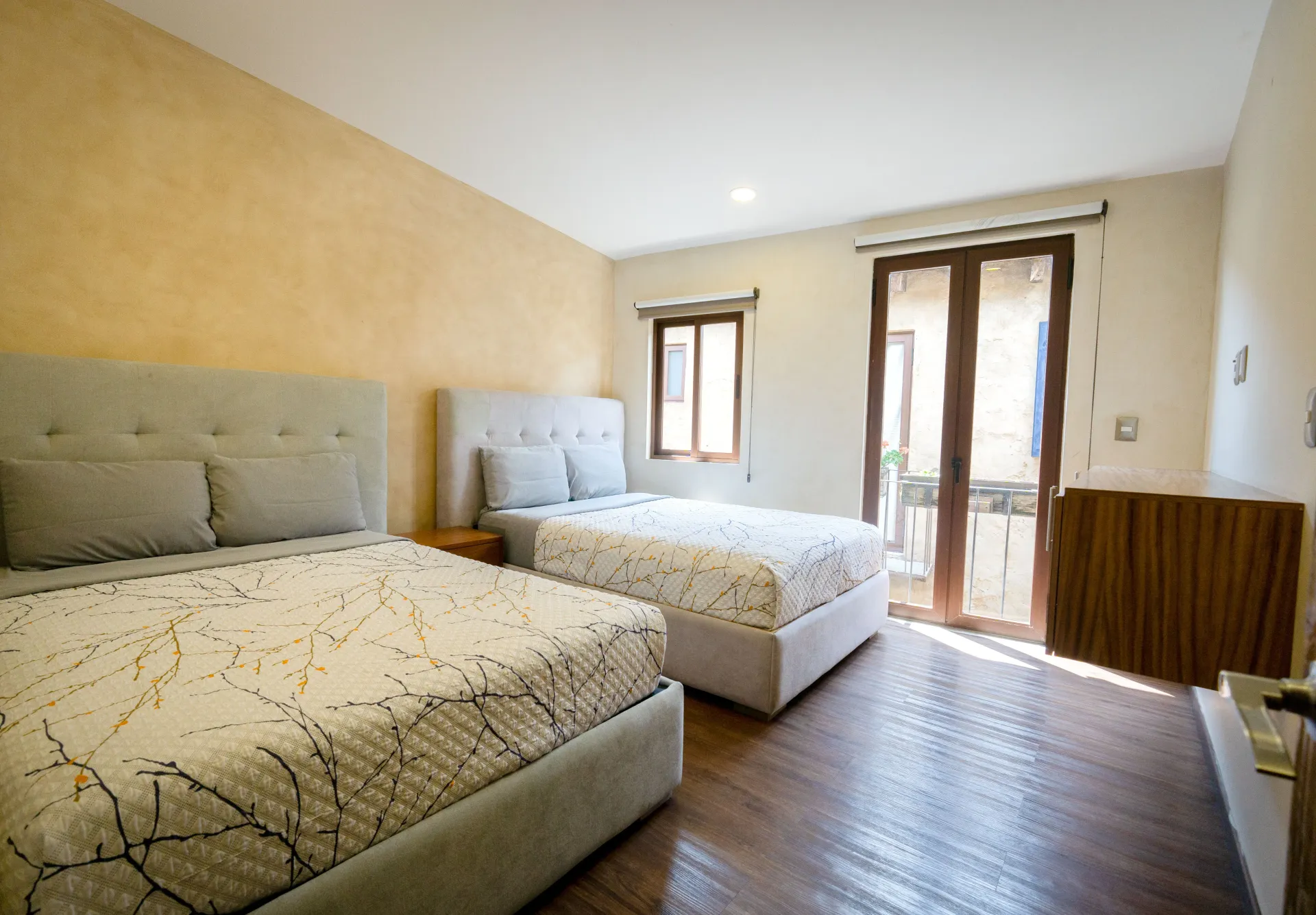 Bedroom with two beds, a small window, and french doors leading to a balcony. Beige and brown tones.