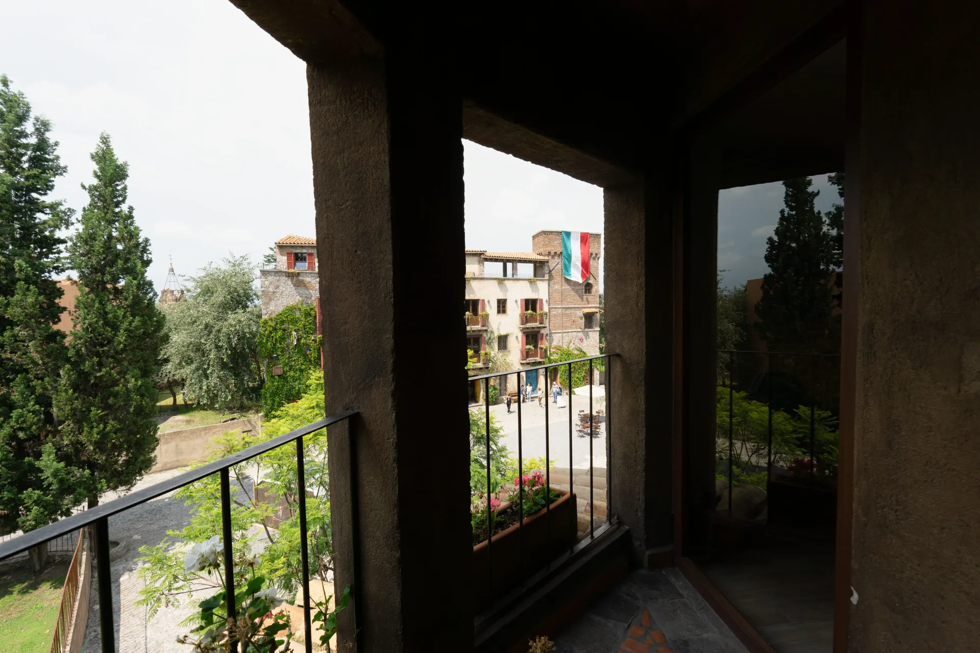 View from a balcony. Dark columns frame a city scene, with trees, buildings, and a colorful flag visible.