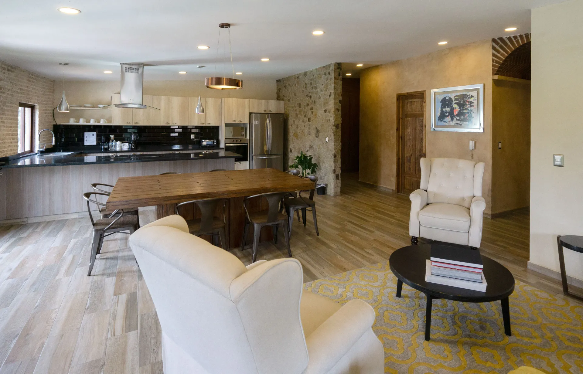 Open-concept kitchen and living area with wood dining table, stone wall, and white armchairs.