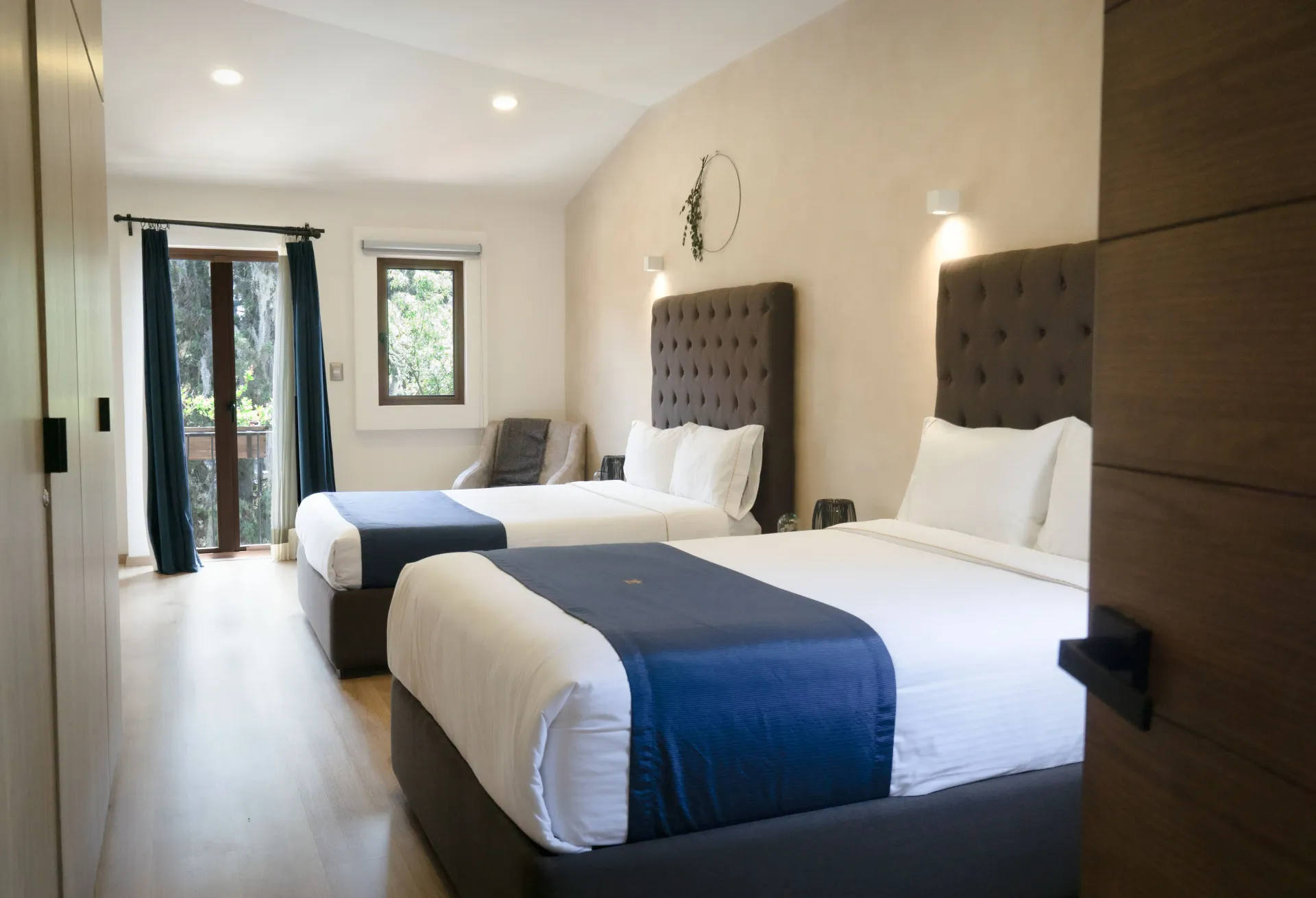 Two beds with white linens and blue accents in a bright room with a doorway to a balcony.