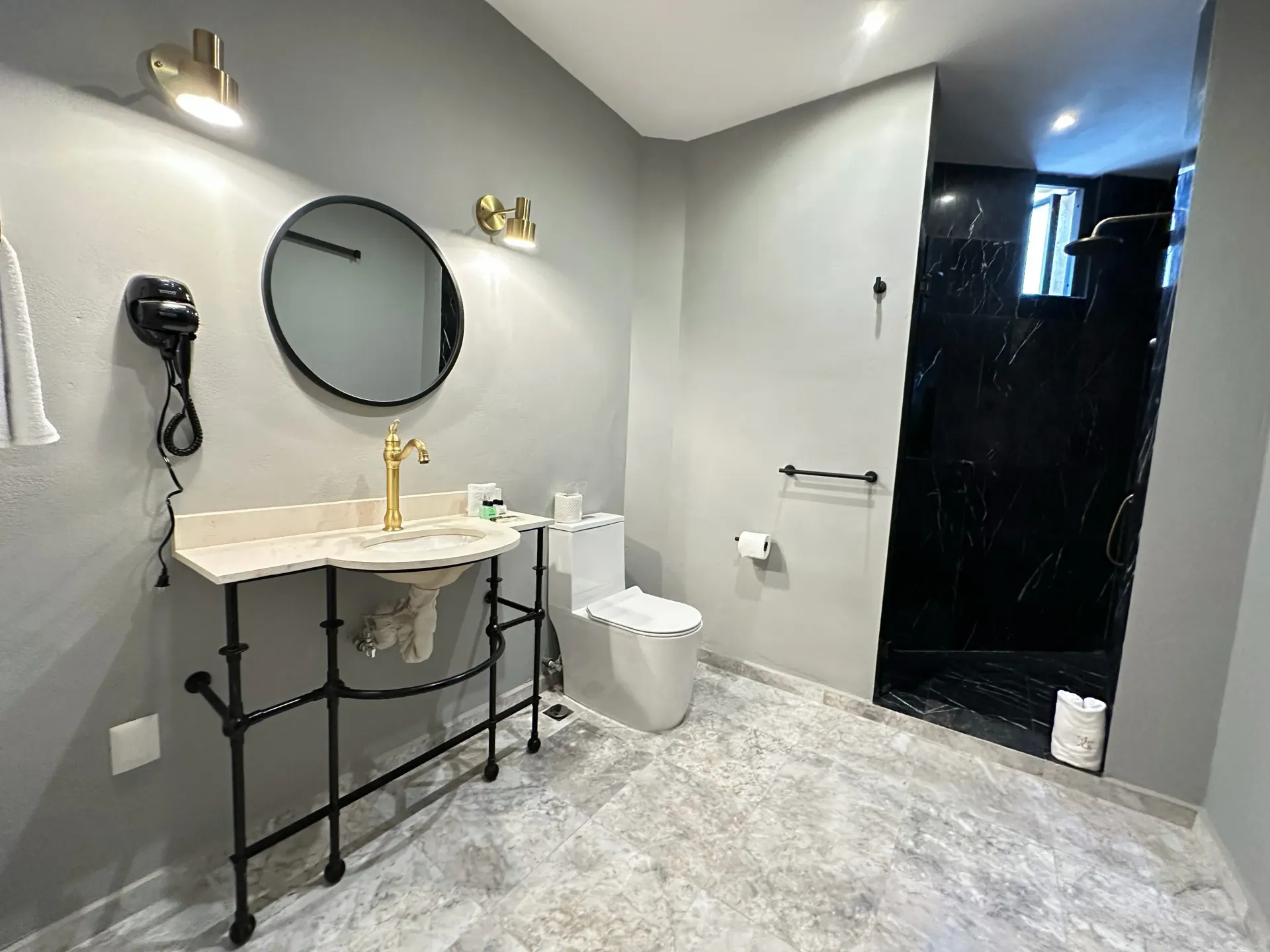 Bathroom with gray walls and floor, marble sink, gold faucet, black-tiled shower, and round mirror.