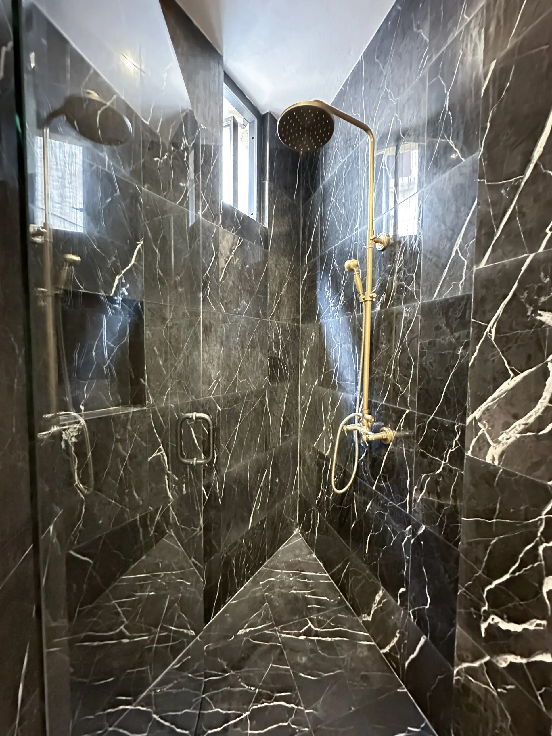 Black marble shower with gold fixtures, including a rain shower head. Two small windows.