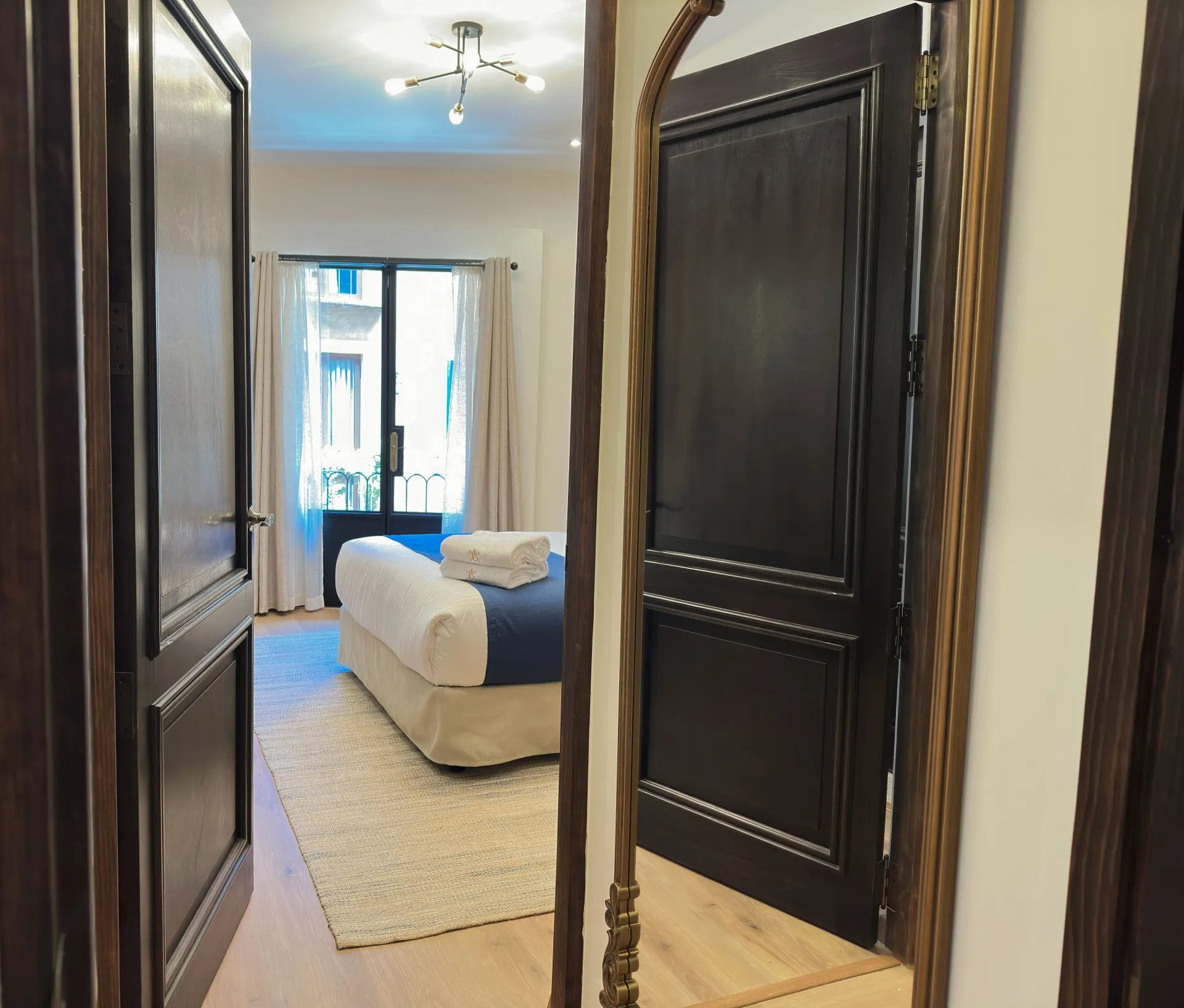 Bedroom view through doorways, dark wood, bed with blue blanket, rug, open doors to balcony.