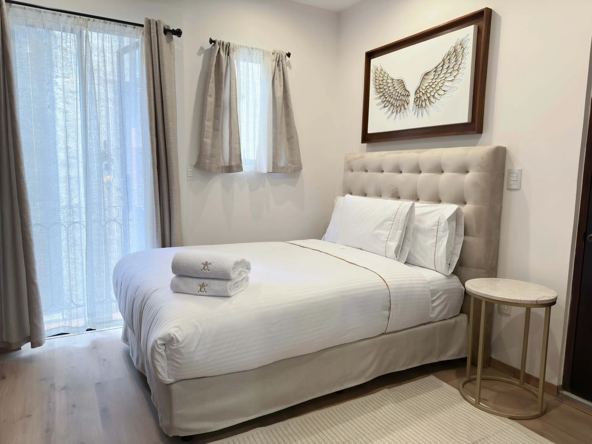 Bedroom with a bed, side table, and artwork. Neutral colors, curtains, and natural light.