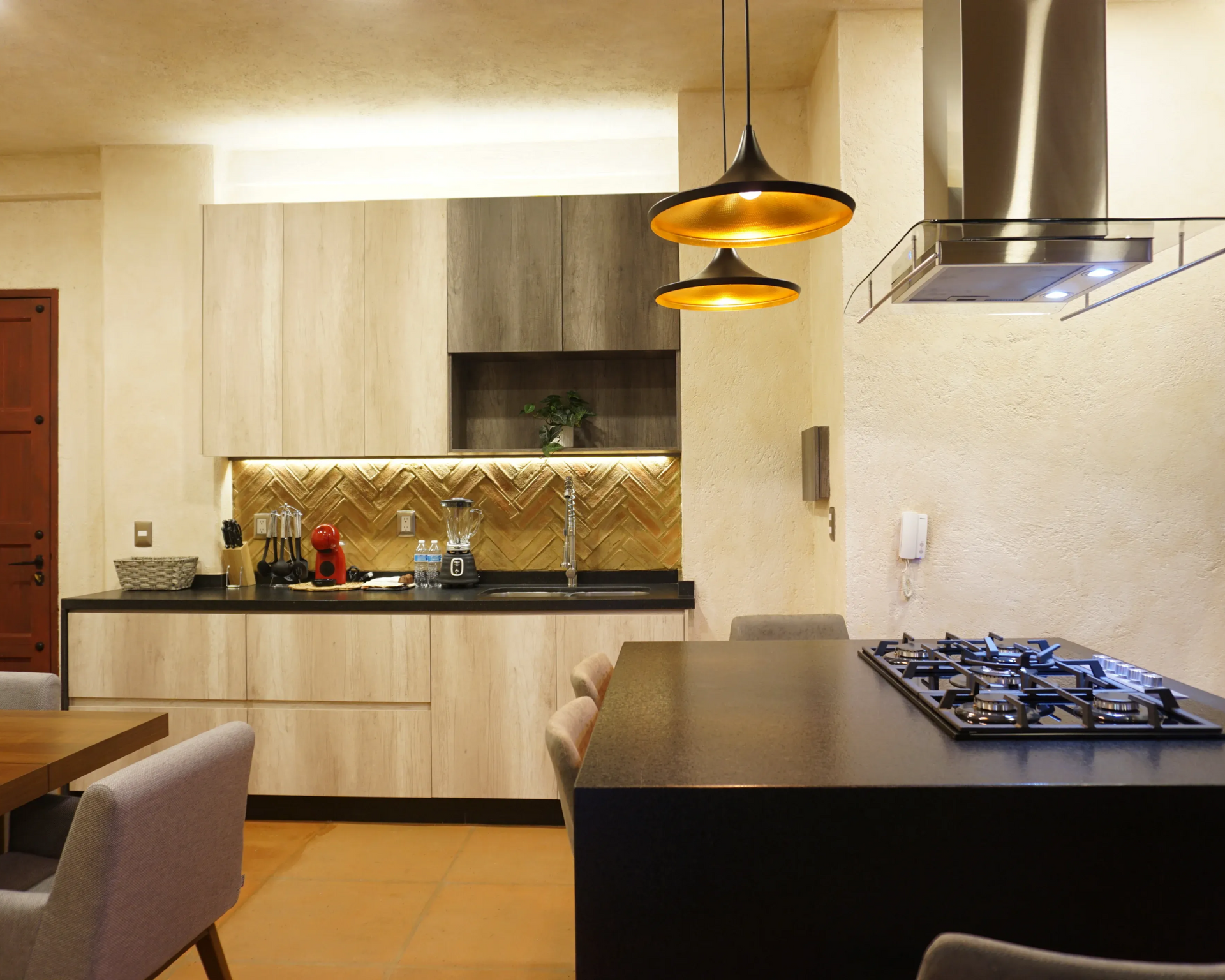Cocina moderna con isla y estufa, gabinetes de madera, protector contra salpicaduras dorado y luces colgantes.