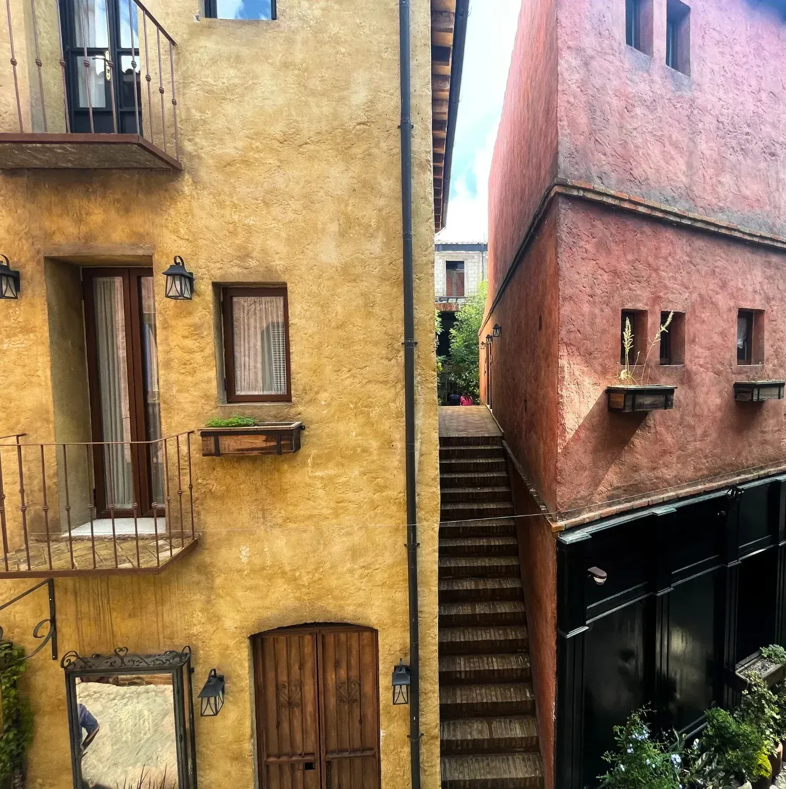 Yellow and red buildings flank a narrow passage with outdoor stairs. A mirror hangs on the yellow building.