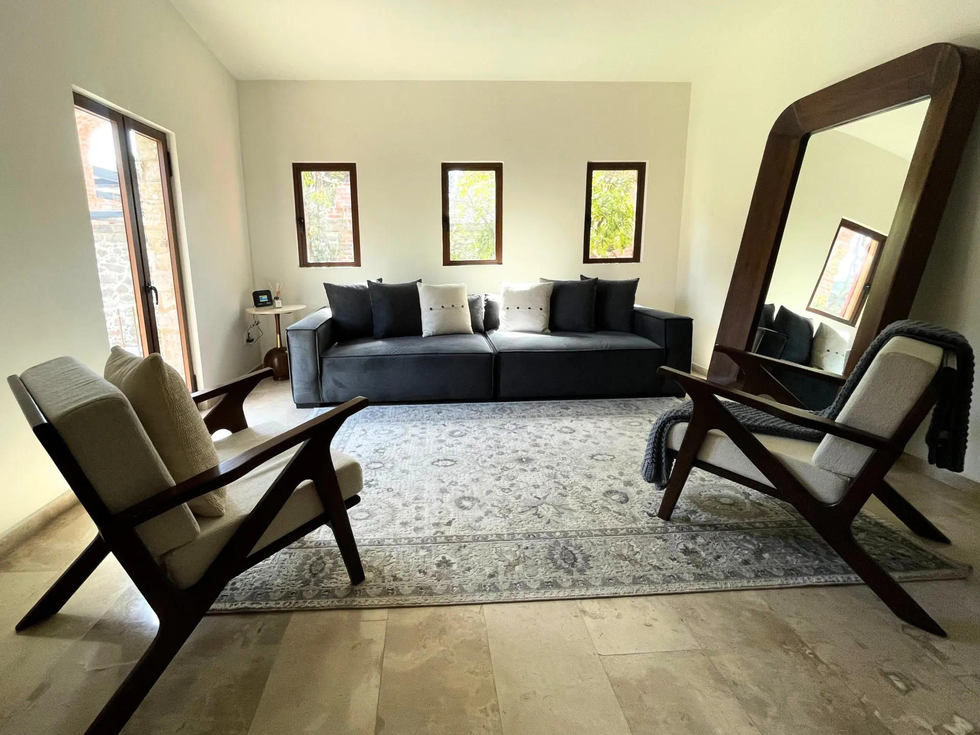 Living room: dark gray sofa, two chairs, large mirror, small windows, and a rug.