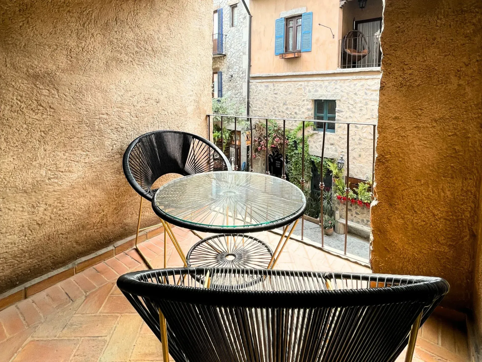 Balcón con sillas y mesa de mimbre negro, con vista a un callejón de una ciudad europea.