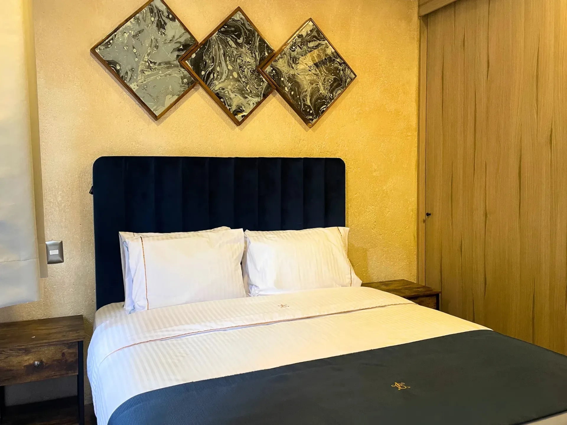 Dormitorio con cabecero azul, ropa de cama blanca y arte de pared abstracto.