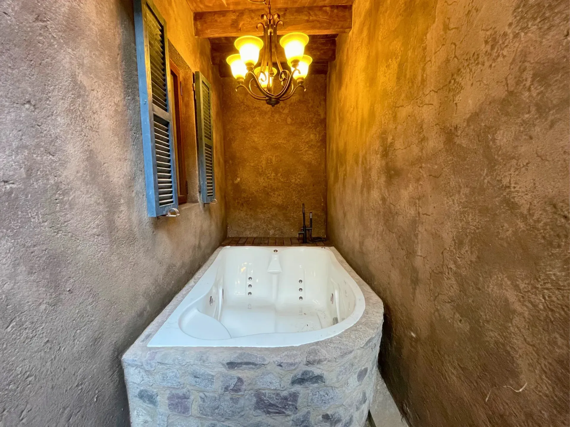 Jacuzzi exterior cerrado en entorno rústico, paredes marrones, lámpara de araña decorativa, pequeña ventana .