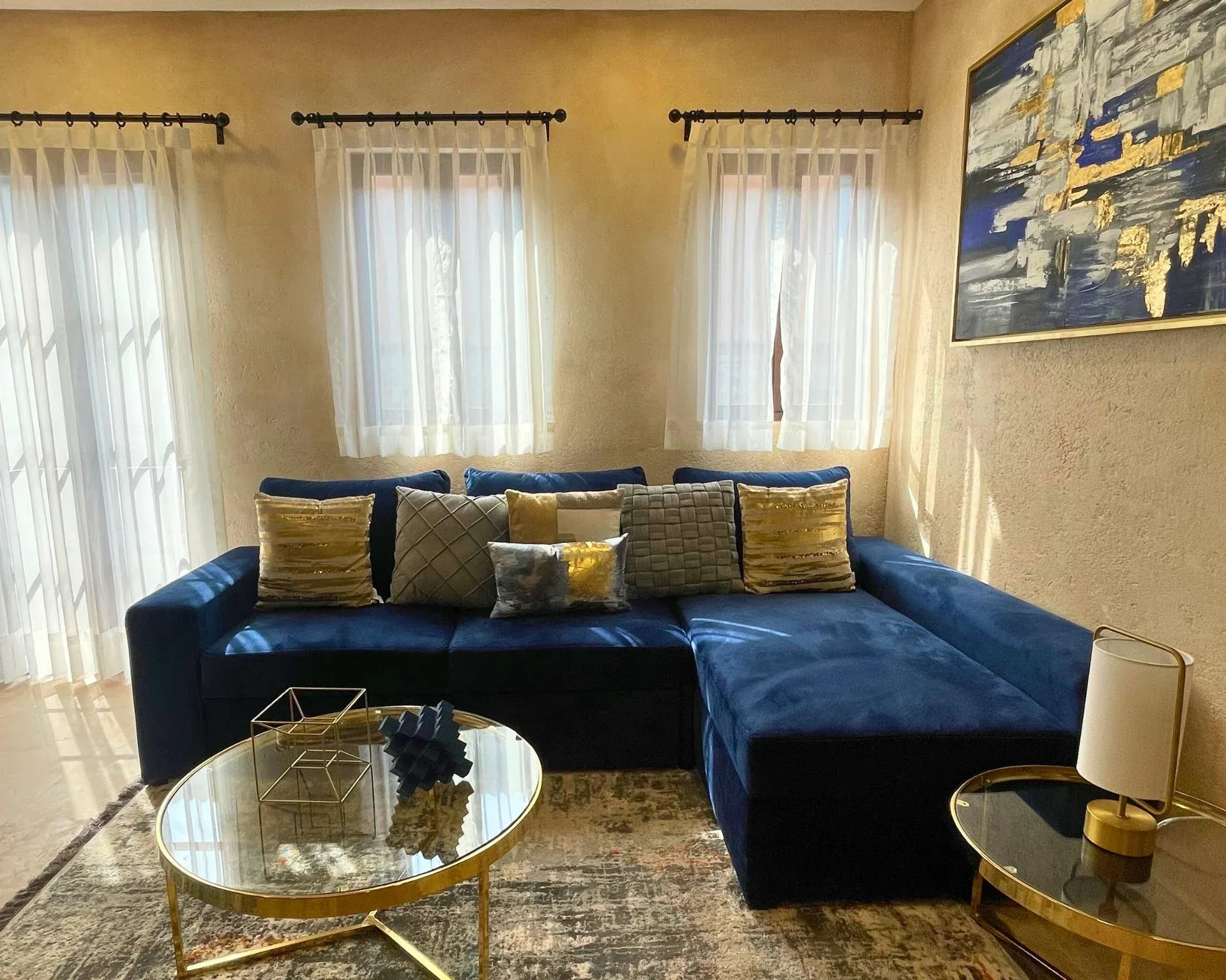 Sofá seccional azul en una habitación con cortinas transparentes, detalles dorados y una pintura moderna.