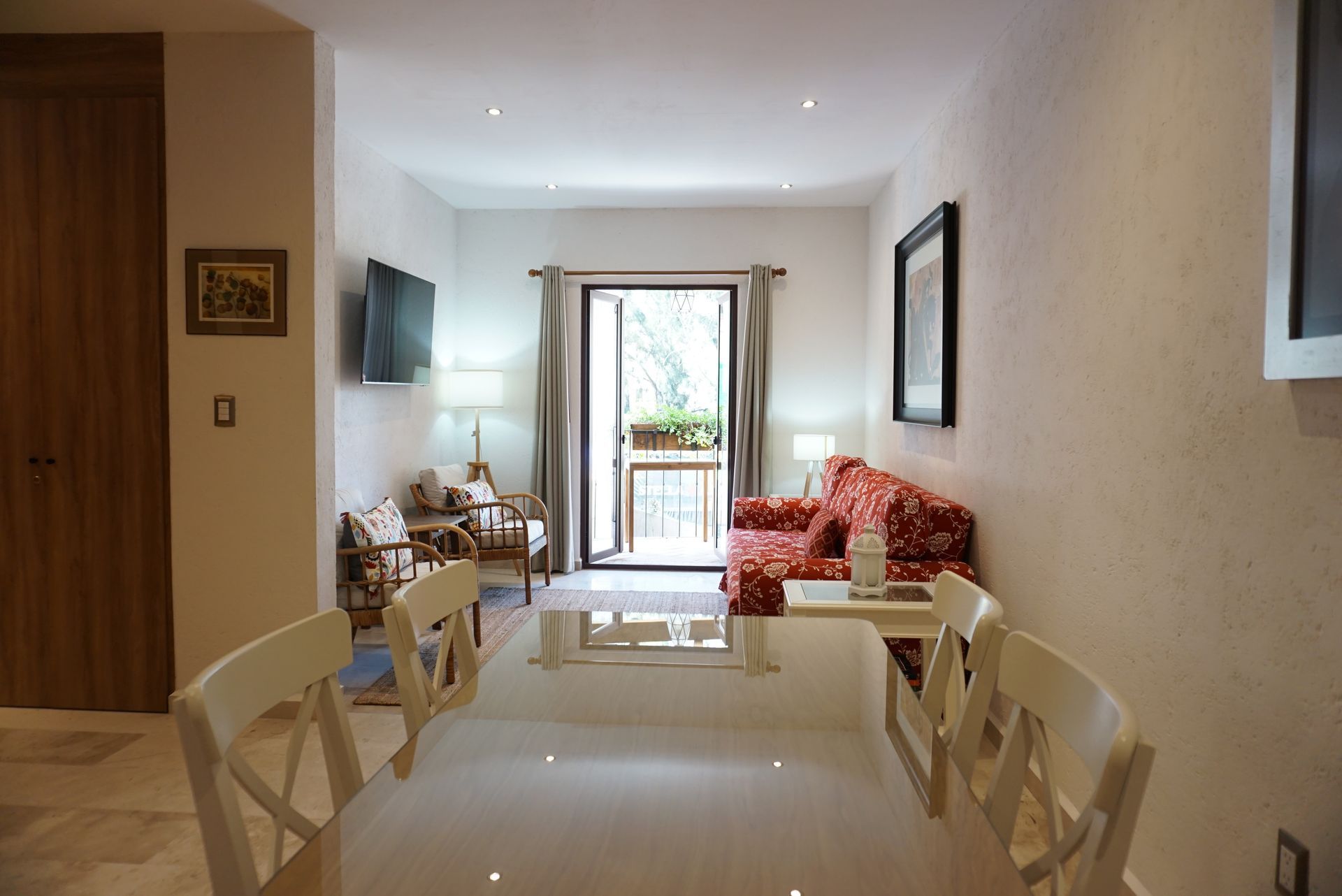 Bright modern living room with a glass dining table, red sofa, and balcony doors at the far end.