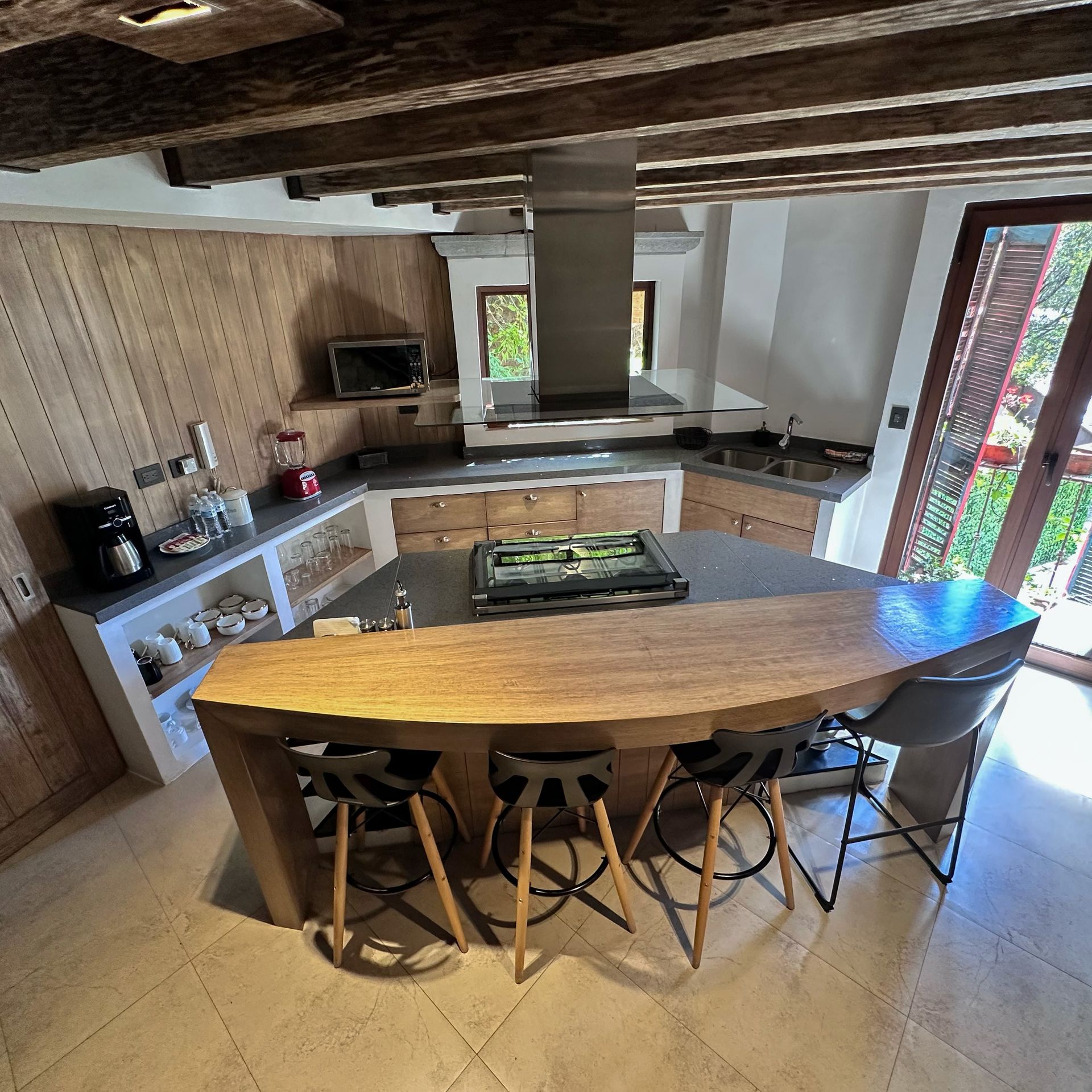 Rustic kitchen with wooden island, stools, stove hood, and large window overlooking greenery