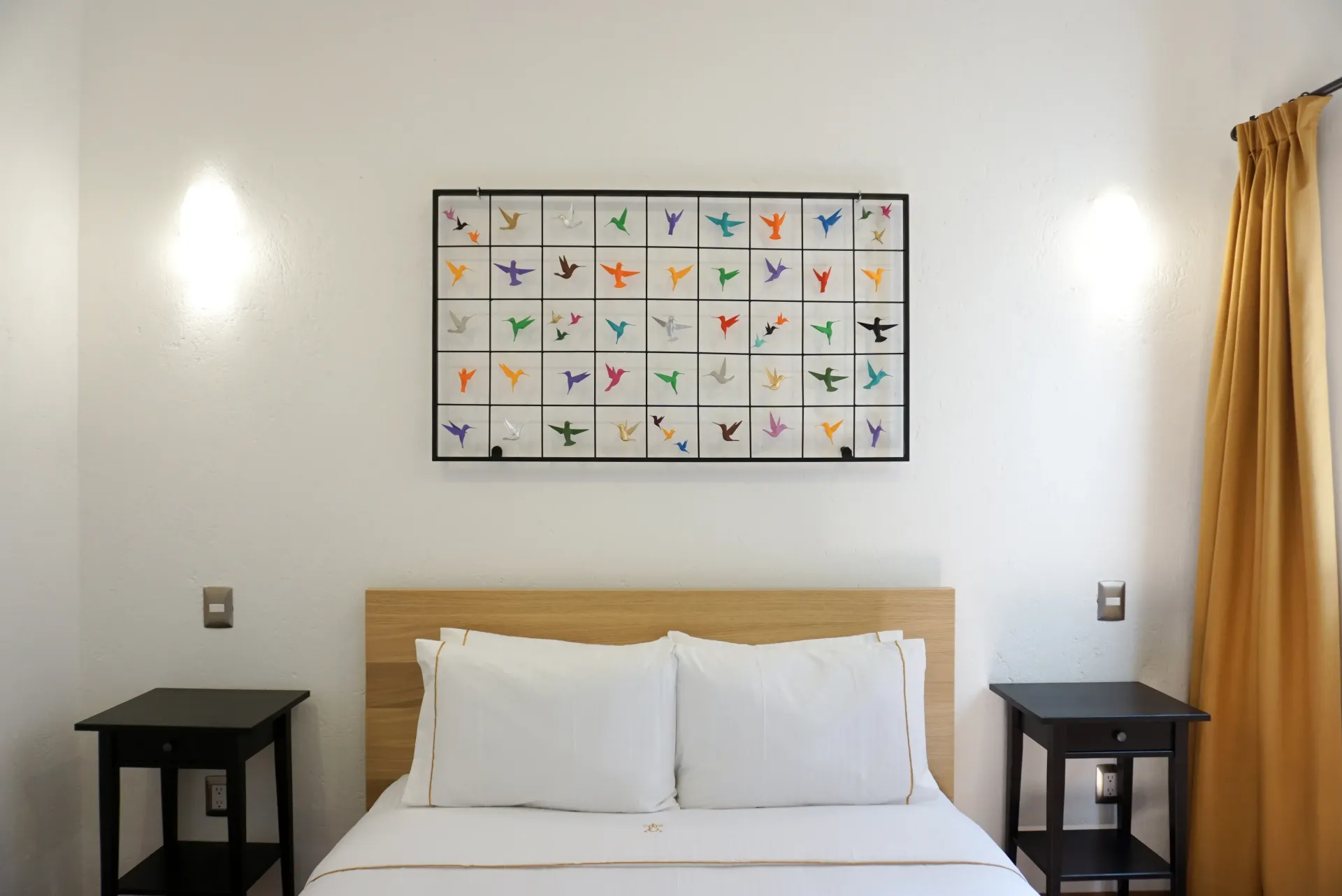 Bedroom with white bedding, artwork of colorful shapes, black nightstands, and gold curtain.