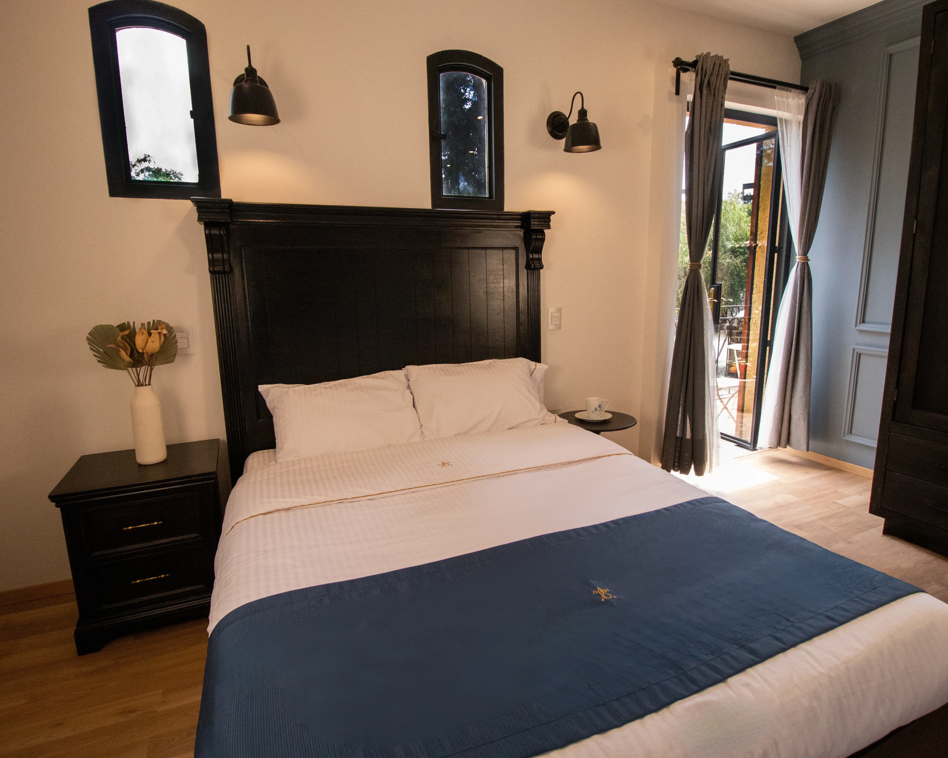 Dormitorio con cabecero negro, colcha azul, mesita de noche, ventana y puerta abierta a un patio.