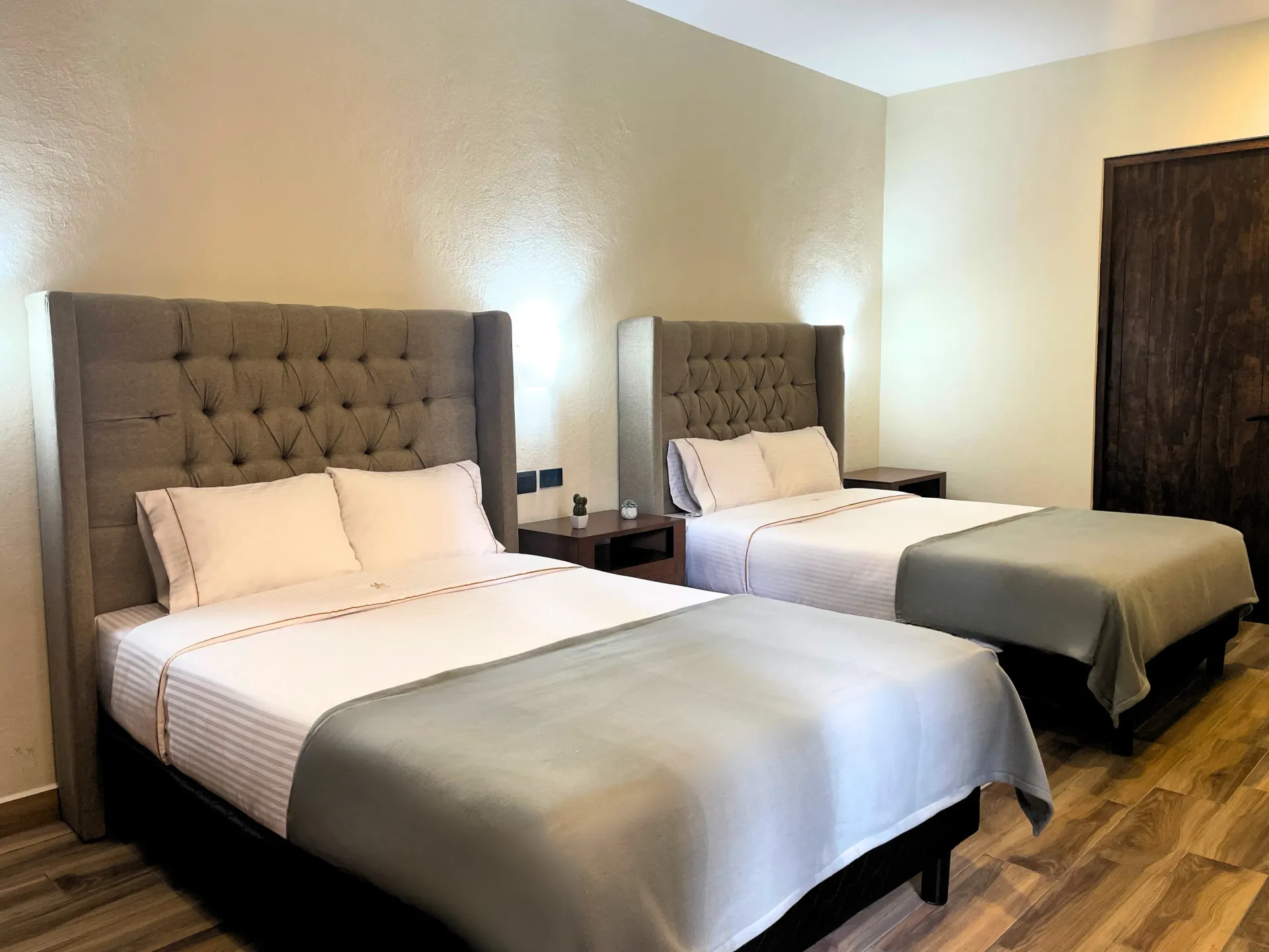 Two beds in a hotel room with gray headboards and white bedding. Wooden floors and a brown door.