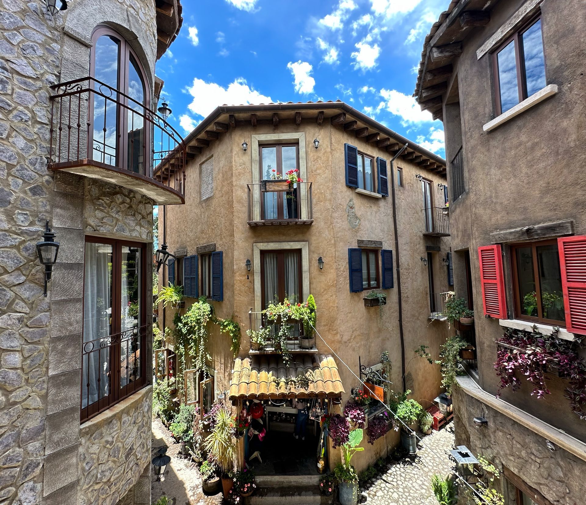 Stone buildings with balconies and colorful shutters, flowers, and a blue sky.