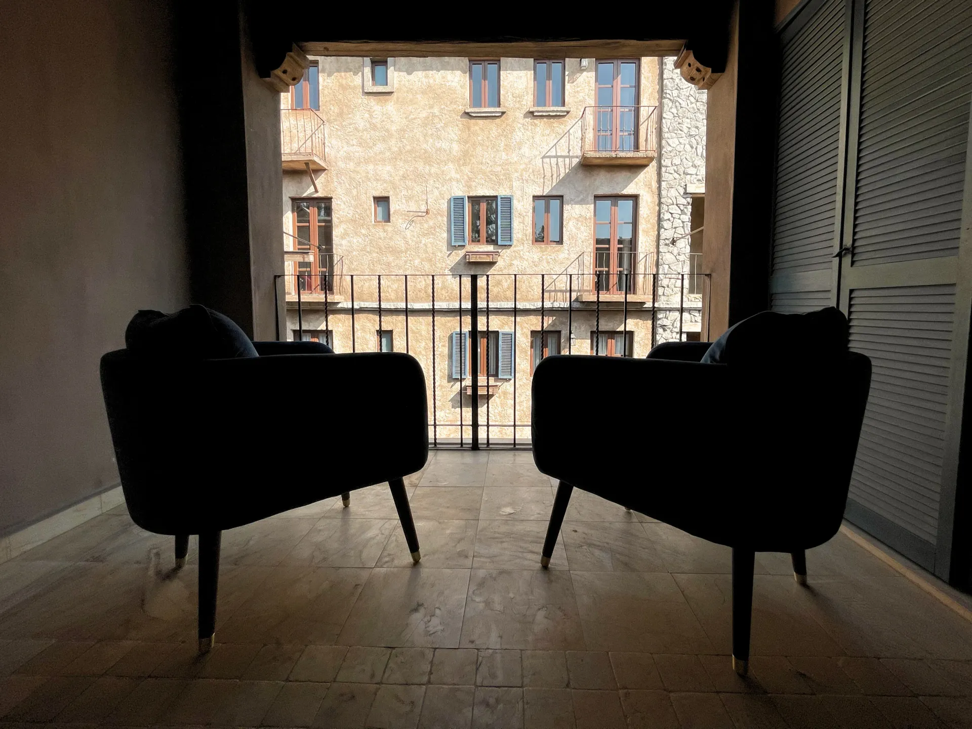 Two dark armchairs on a balcony face a sunlit building with windows.