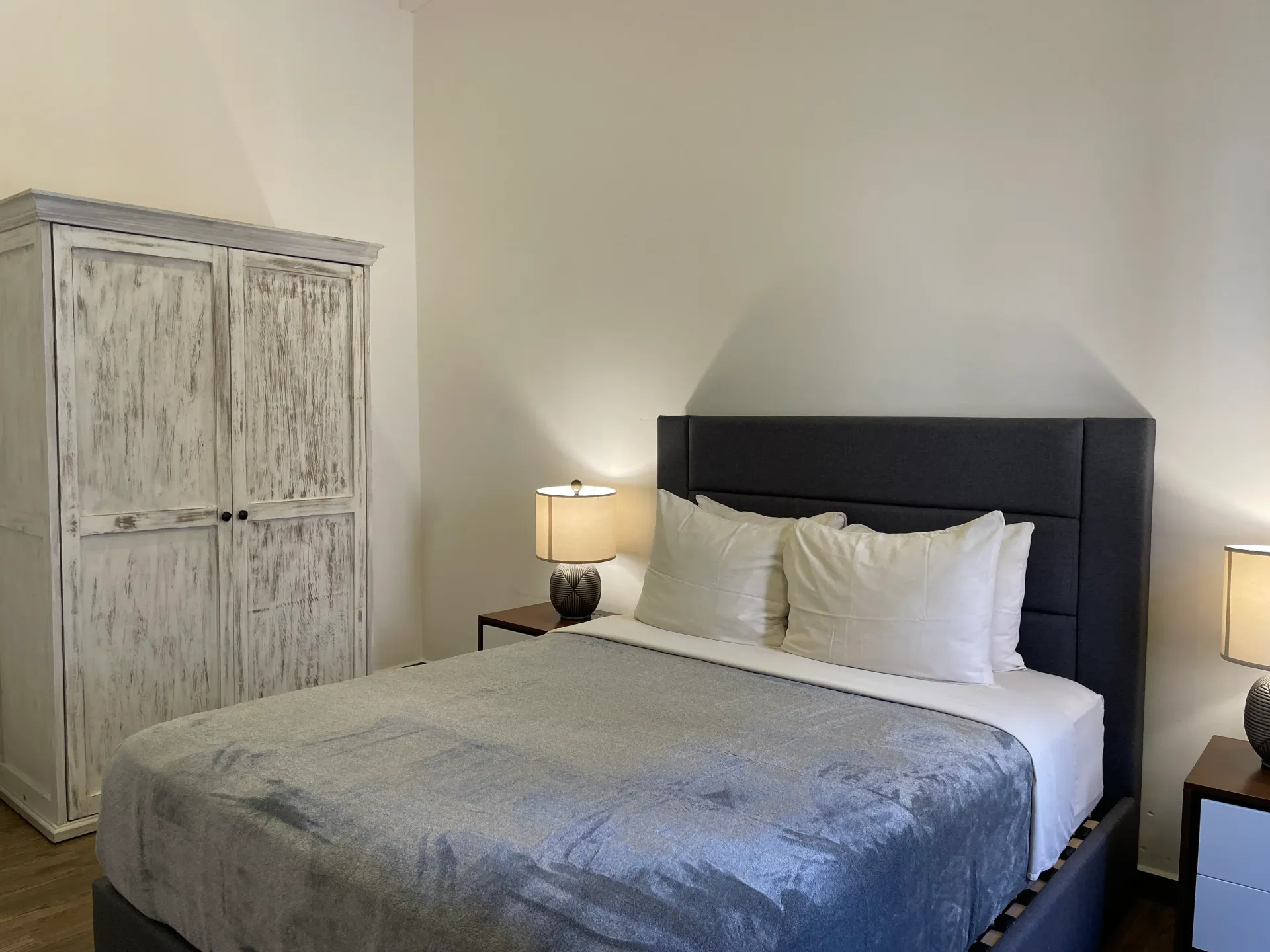 Bedroom with a bed, nightstands, lamps, and a distressed white wardrobe.