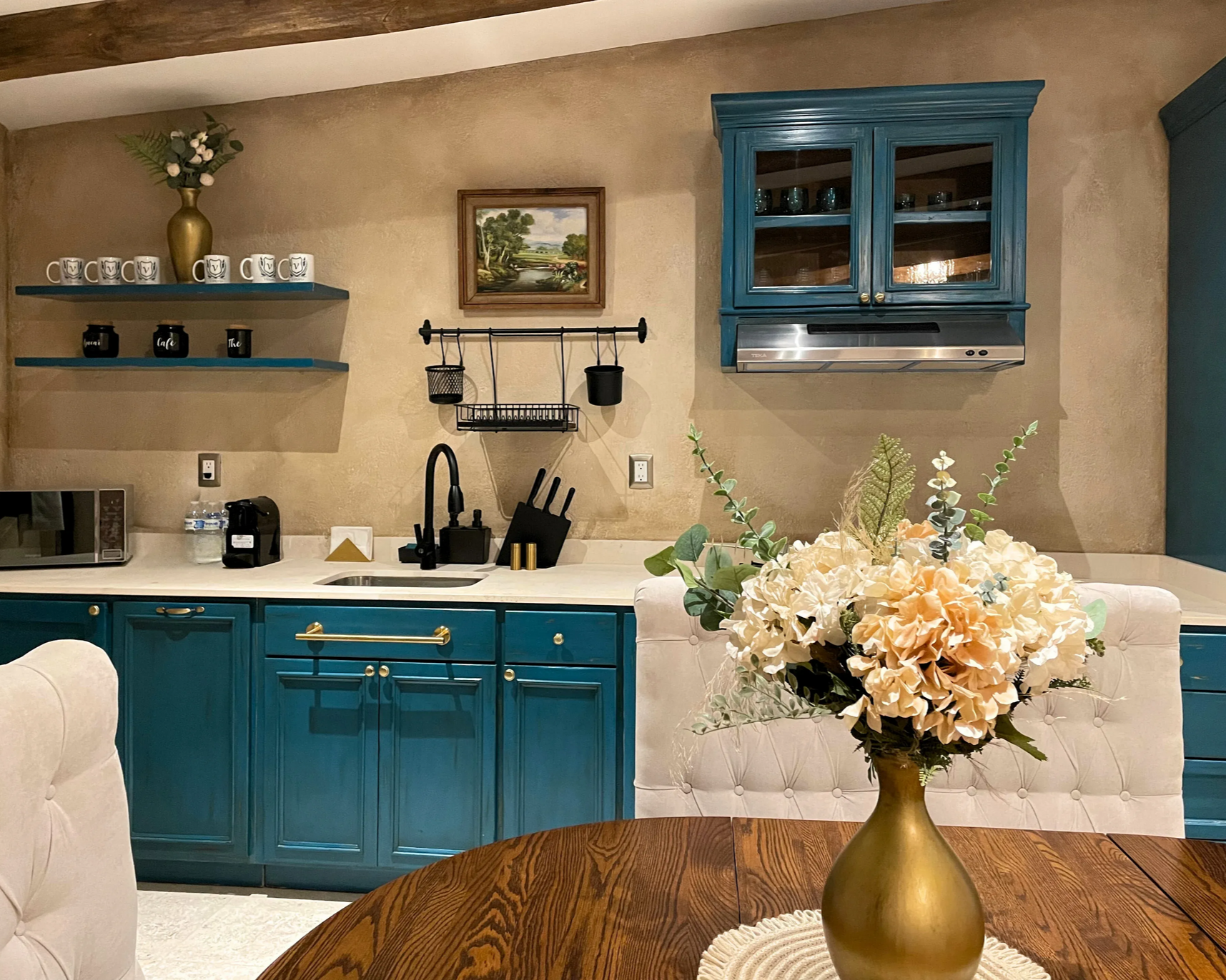 Cocina verde azulado con mesa de madera, encimera blanca y arreglo floral.