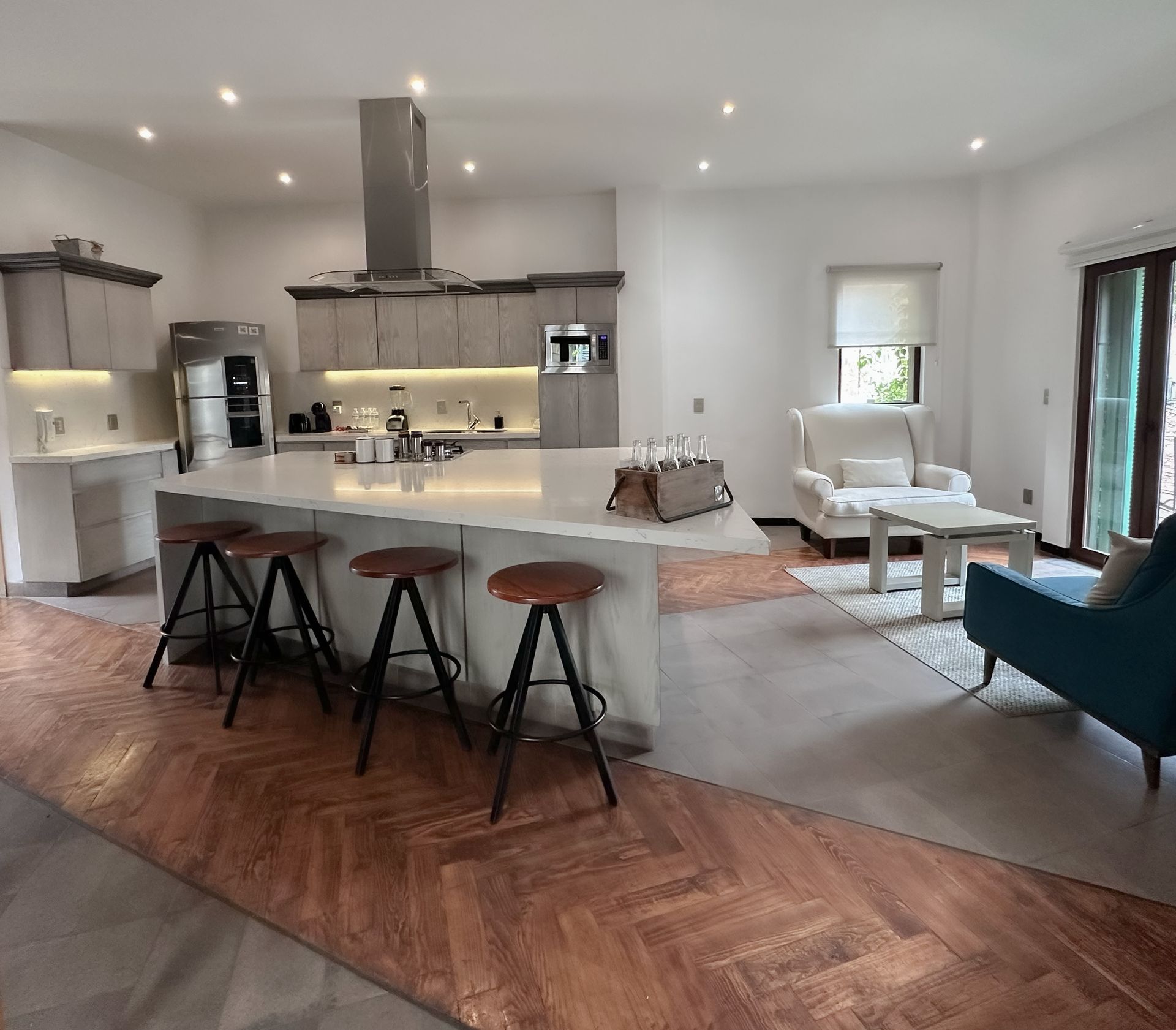 Modern open-plan kitchen and living room with island stools, wood floors, white furniture, and a blue chair