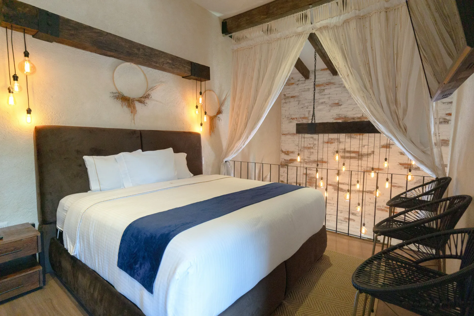 Bedroom with bed, hanging lights, chairs, and draped curtains, rustic decor with neutral tones.
