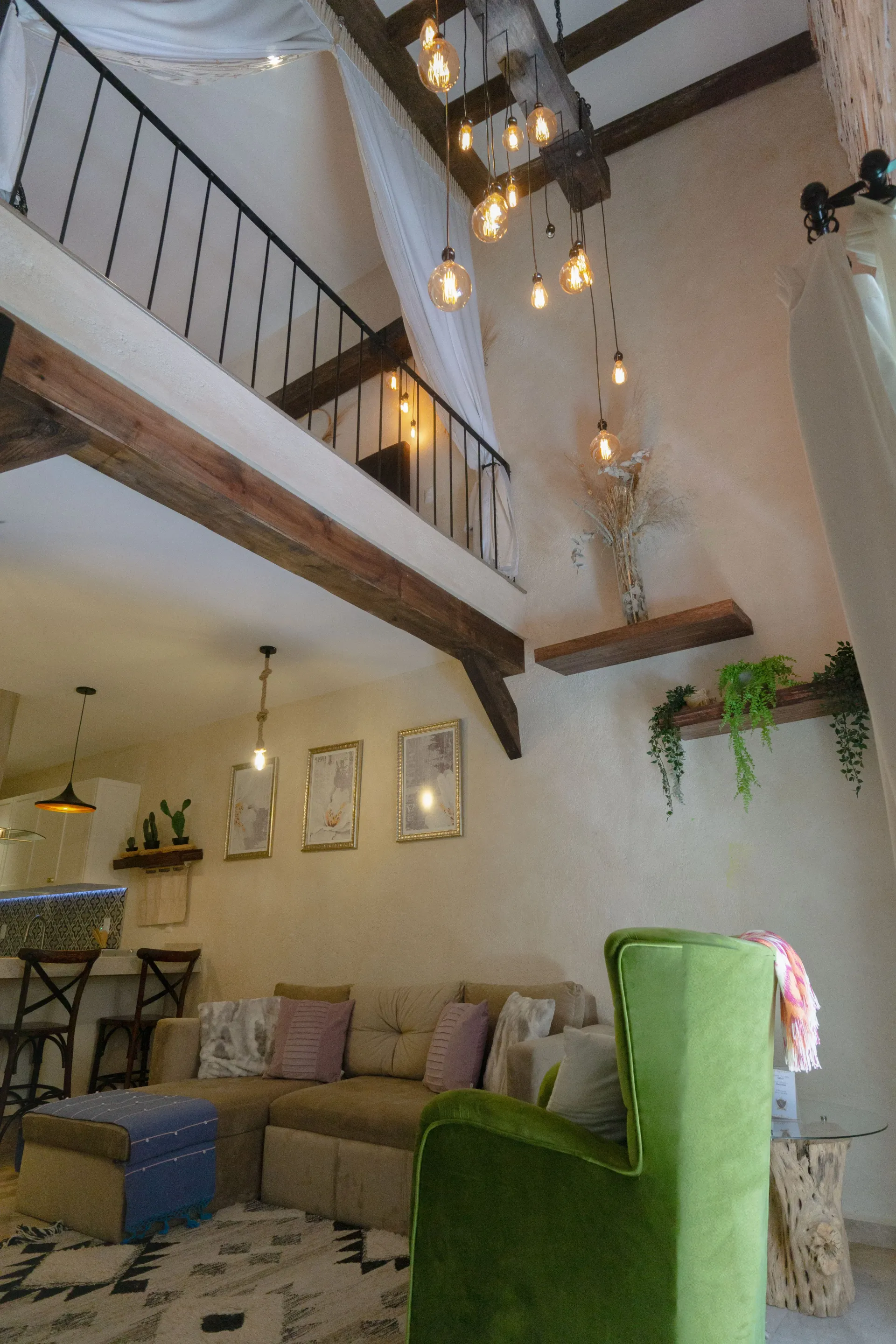 Cozy living room with a loft. Green armchair, neutral sofa, and exposed wooden beams. Decorative lights and plants.