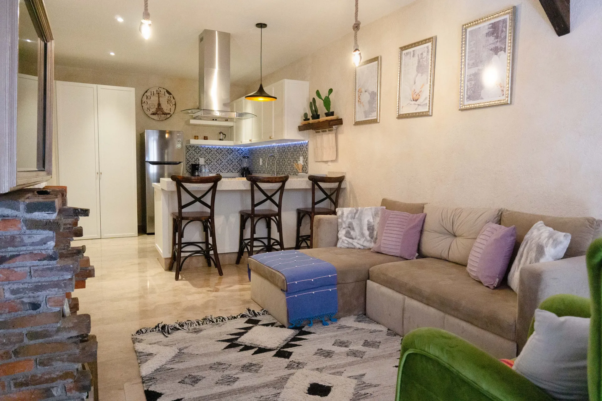 Cozy living room with kitchen. Beige couch, patterned rug, brick accents, bar stools, and framed art.