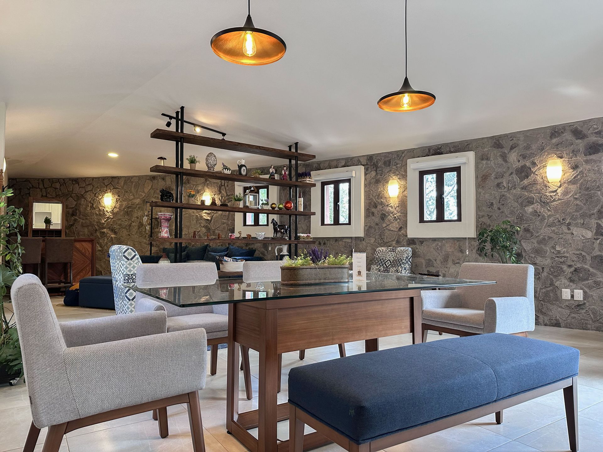 Modern lounge with wicker chairs, blue bench, wooden table, and stone walls under warm pendant lights