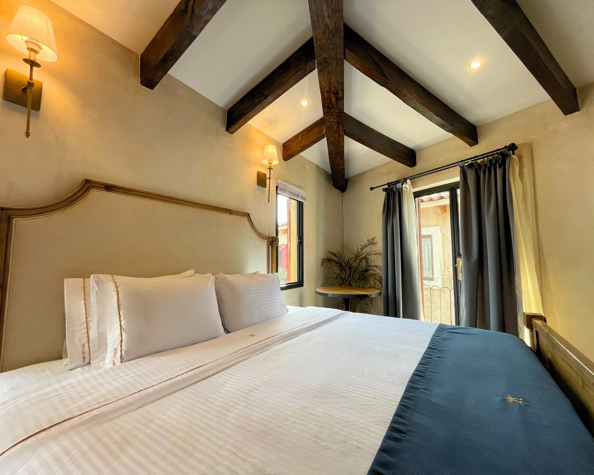 Dormitorio con sábanas blancas, vigas de madera y camino de cama de color azul; ventana con cortinas.