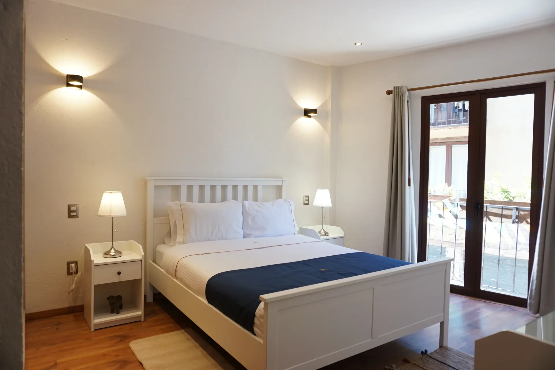 Dormitorio blanco con cama, mesitas de noche, lámparas y puertas a un balcón.