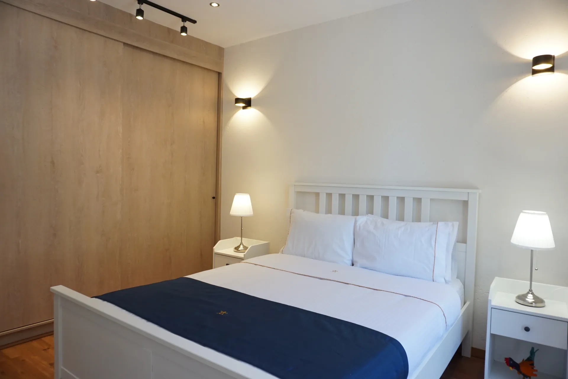 Dormitorio con cama blanca, mesitas de noche y armario de madera. Iluminación cálida.