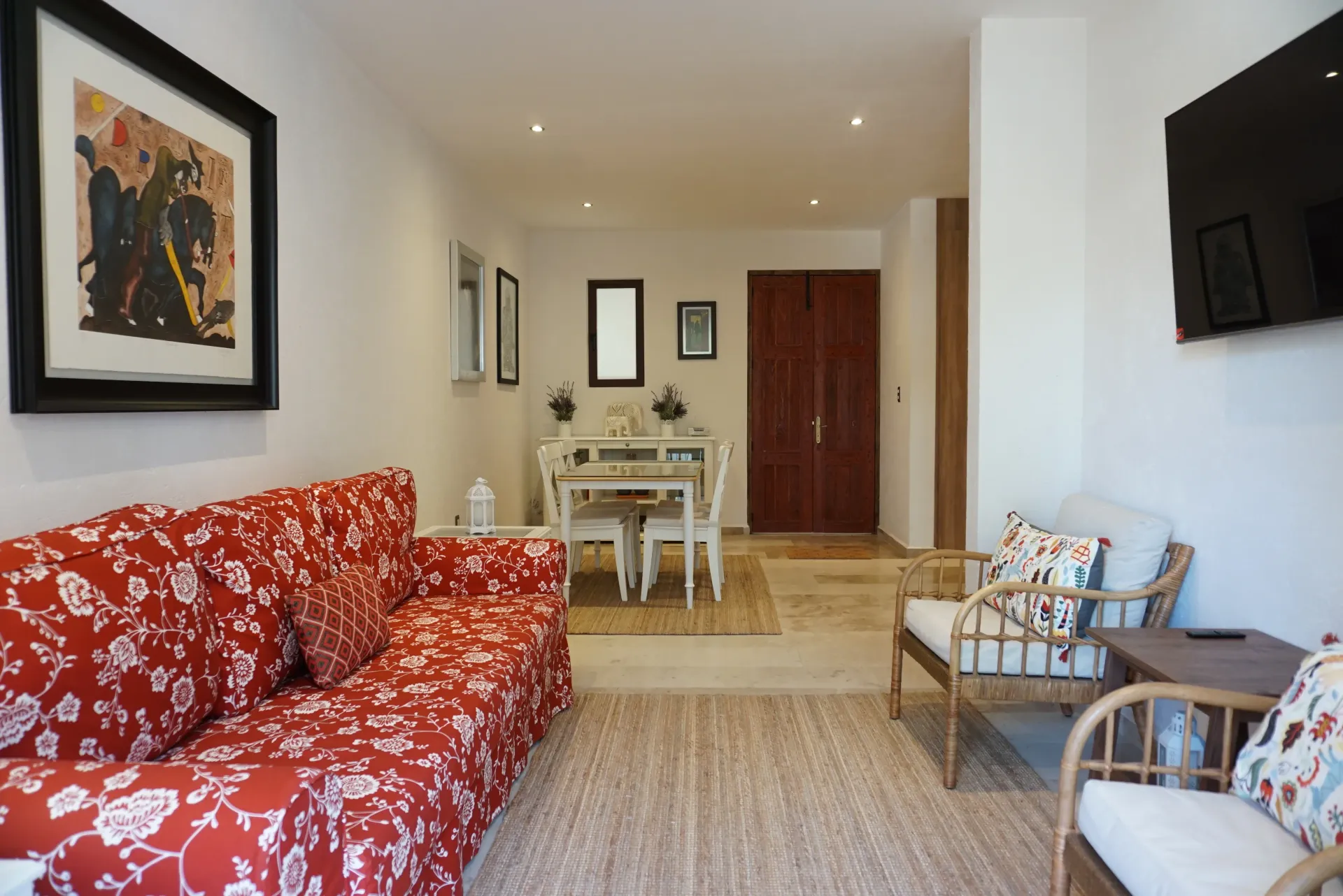 Sala de estar con sofá floral rojo, sillas de ratán, mesa de comedor y obras de arte.