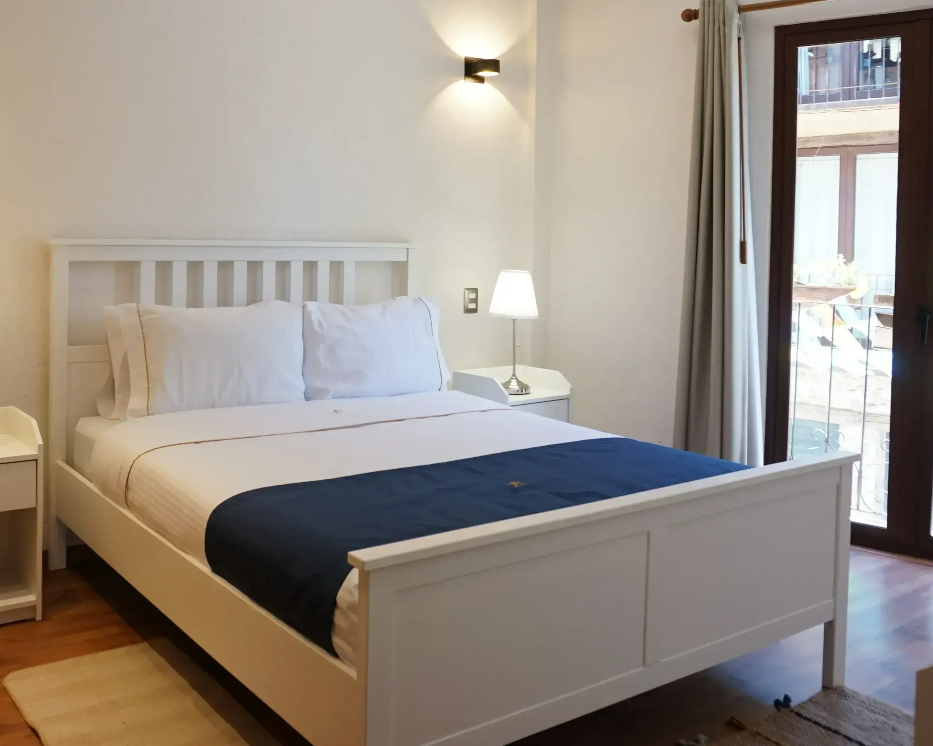 Dormitorio blanco con cama, mesitas de noche y puertas corredizas a un balcón.