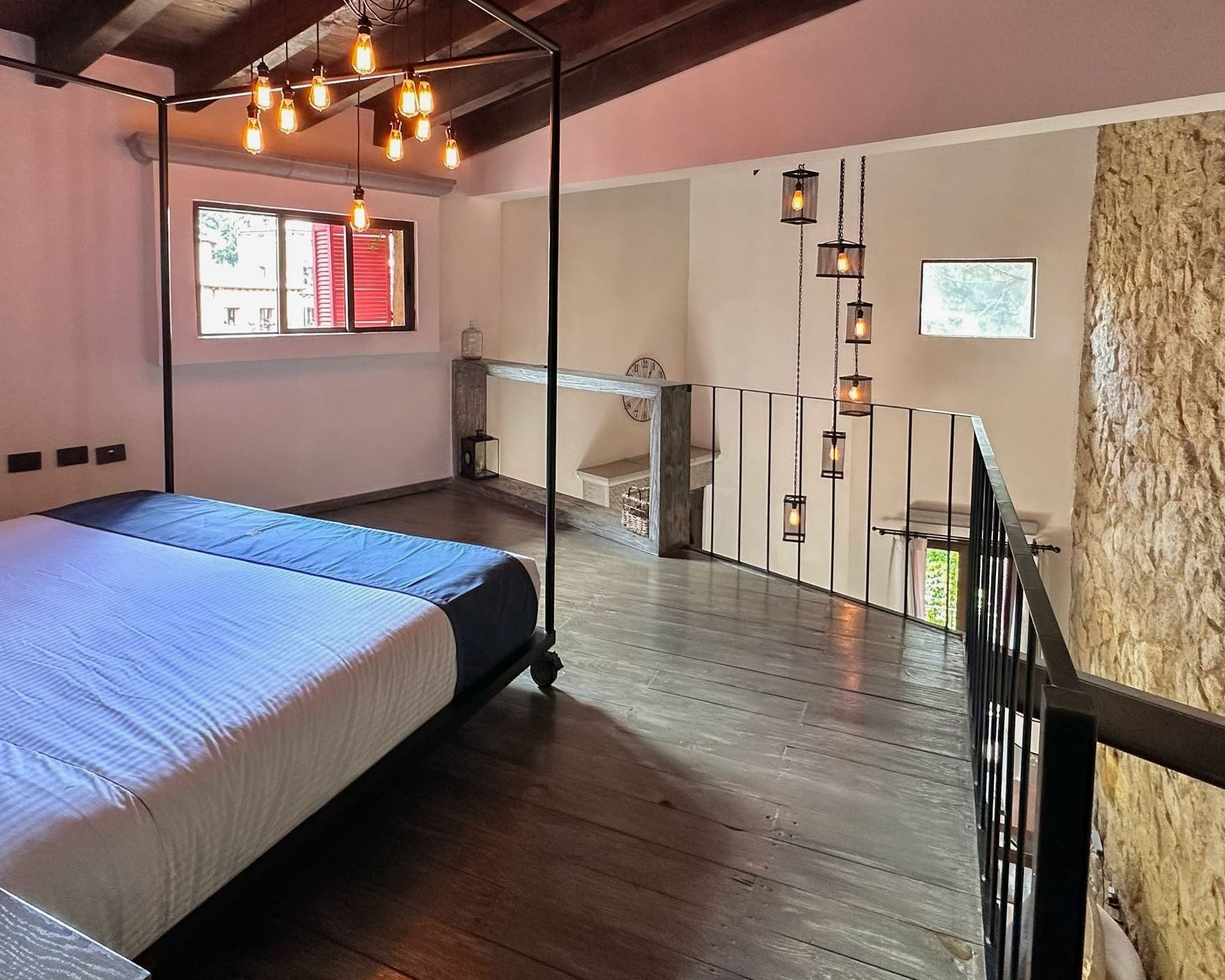 Bedroom with loft, wood floors, dark bed frame, hanging lights, stone wall.