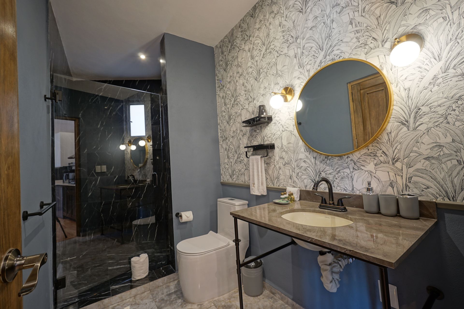 Modern bathroom with gray wallpaper, round mirror, vanity sink, toilet, and glass shower enclosure
