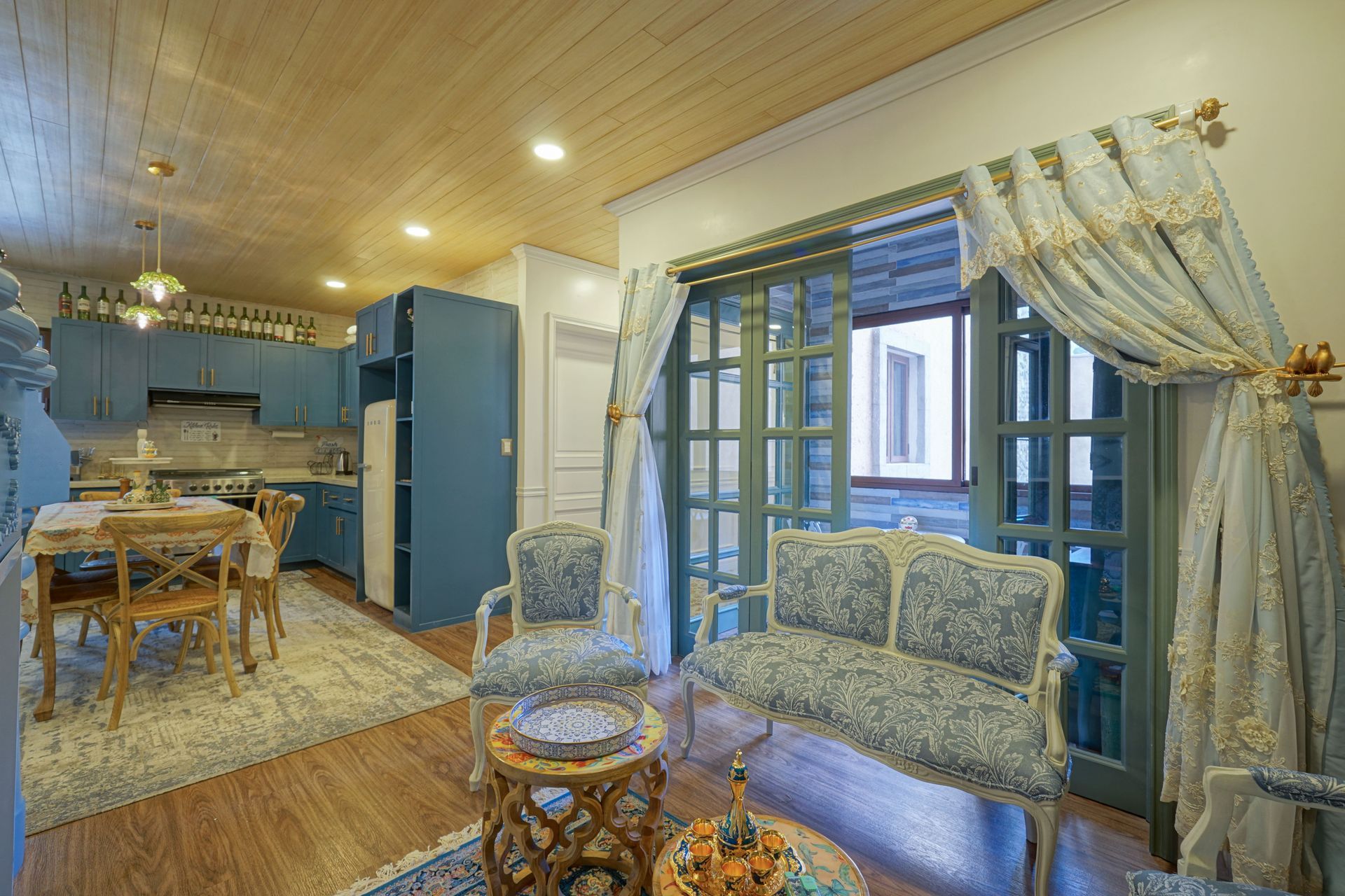 Cozy blue-and-cream living room with sofa, glass table, dining area, and large windows with curtains