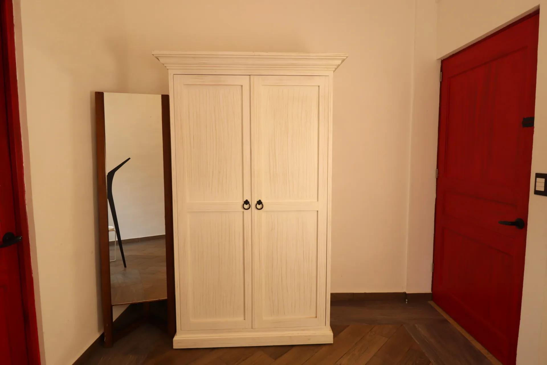 Armario blanco y espejo de cuerpo entero en un pasillo, puertas rojas a cada lado, suelo de madera.