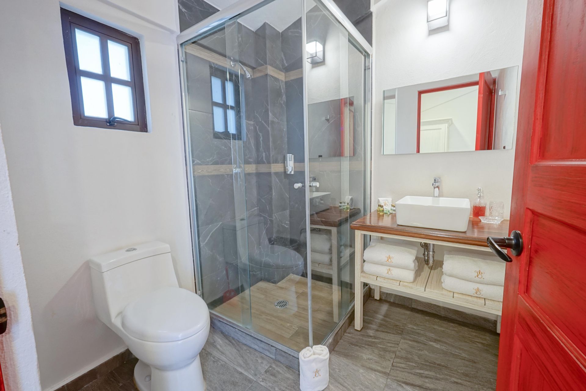 Modern bathroom with glass shower, white toilet, red door, and vanity mirror